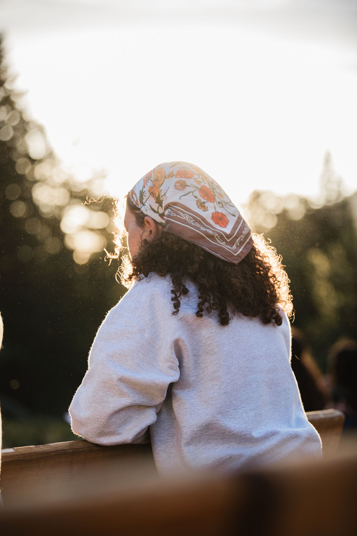 Young girl with curly hair and floral headband in white sweater leaning on wooden fence at golden hour.