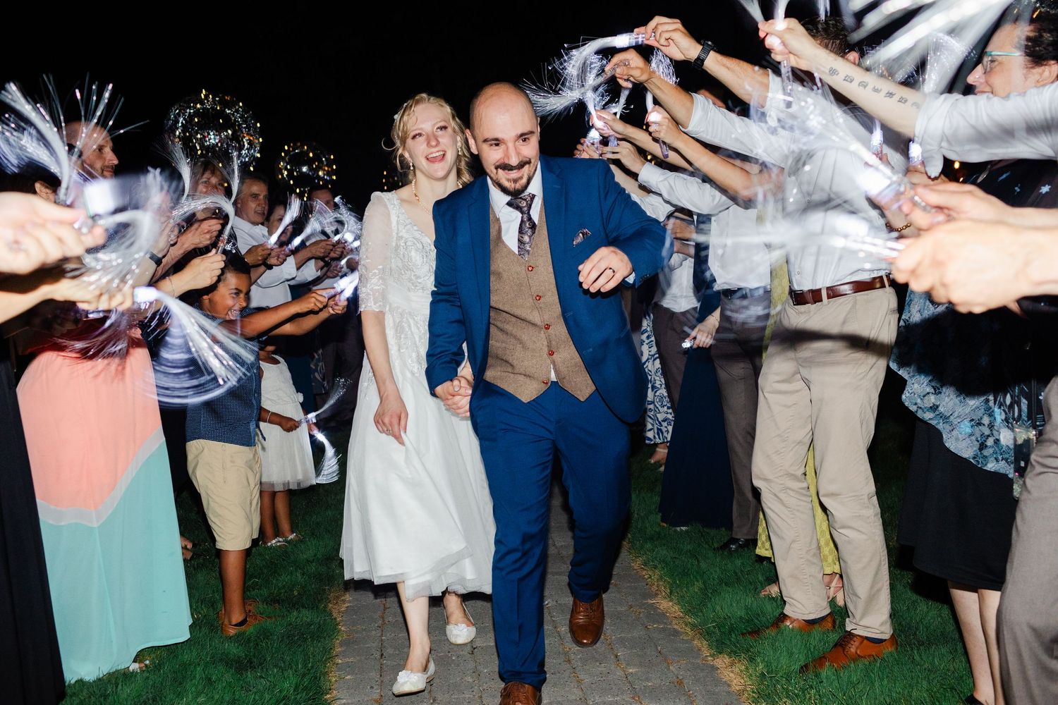 Bride and groom walk through sparkler tunnel at outdoor wedding reception at night, guests cheering around them.