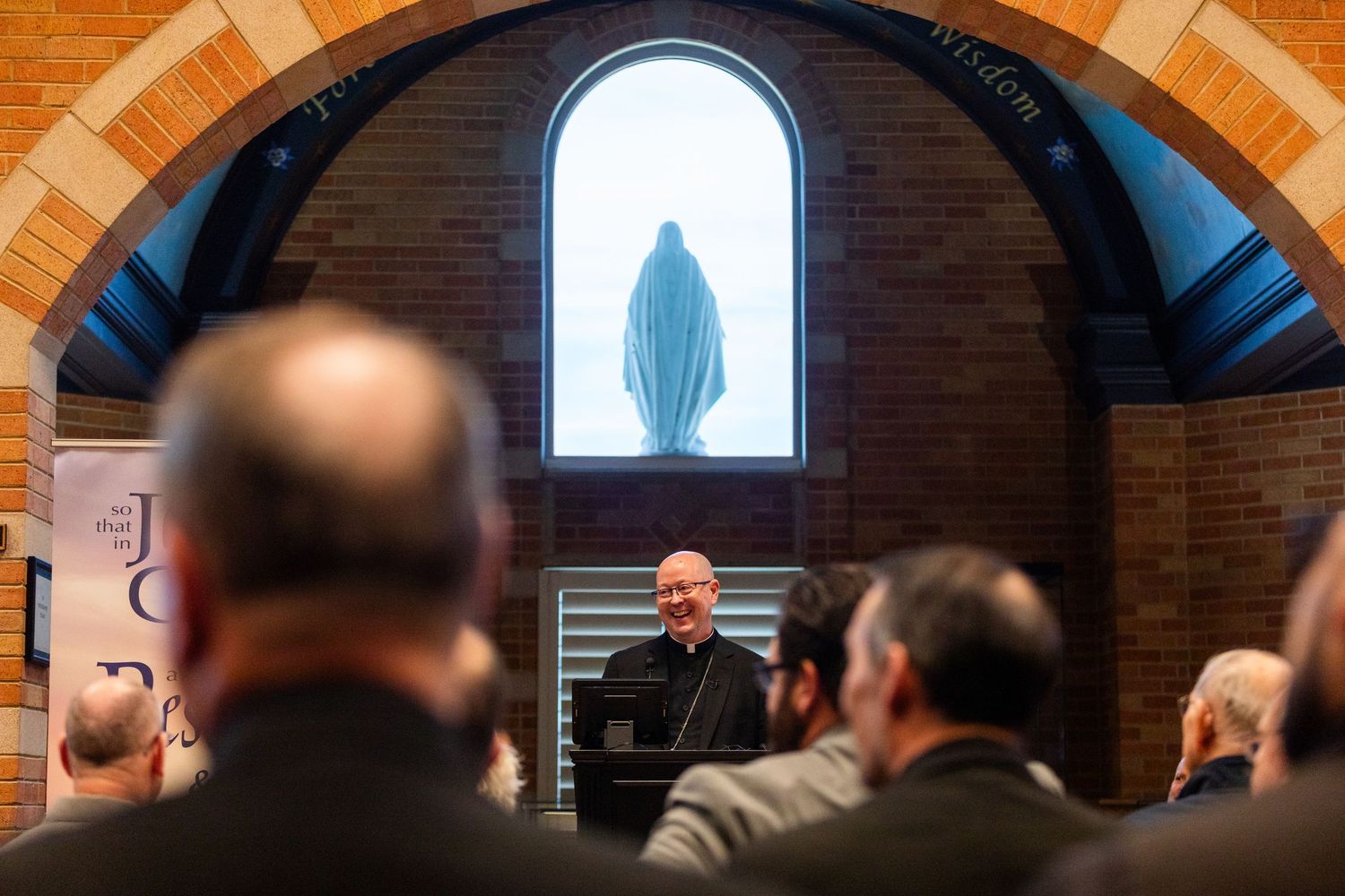 A Catholic priest addresses a gathering inside a brick chapel with an arched ceiling and illuminated Virgin Mary statue.
