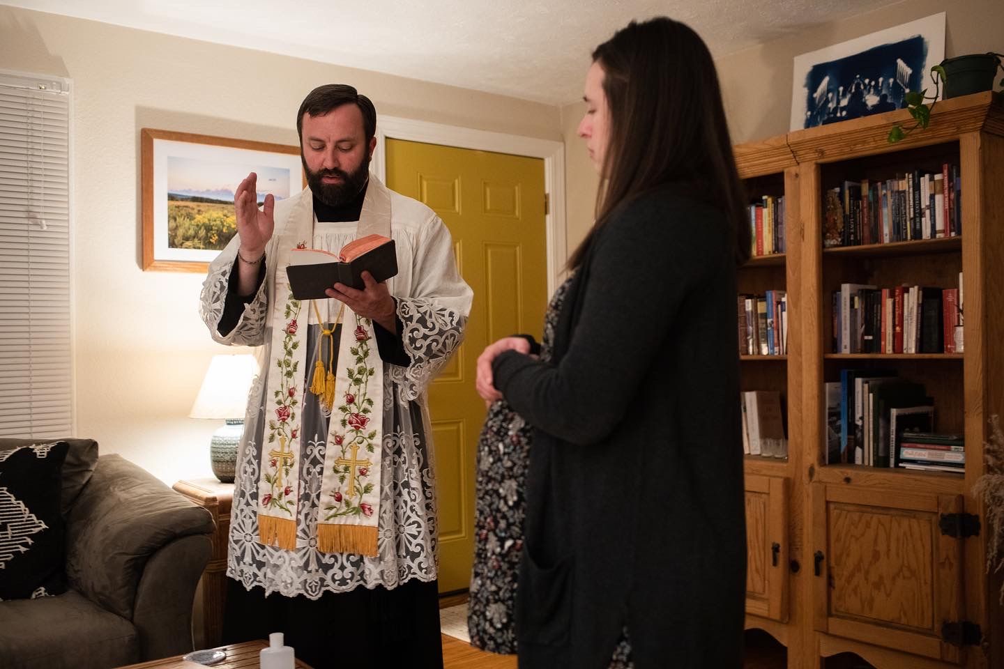 A priest wearing traditional white liturgical vestments with embroidered details speaks to a woman in a home setting.