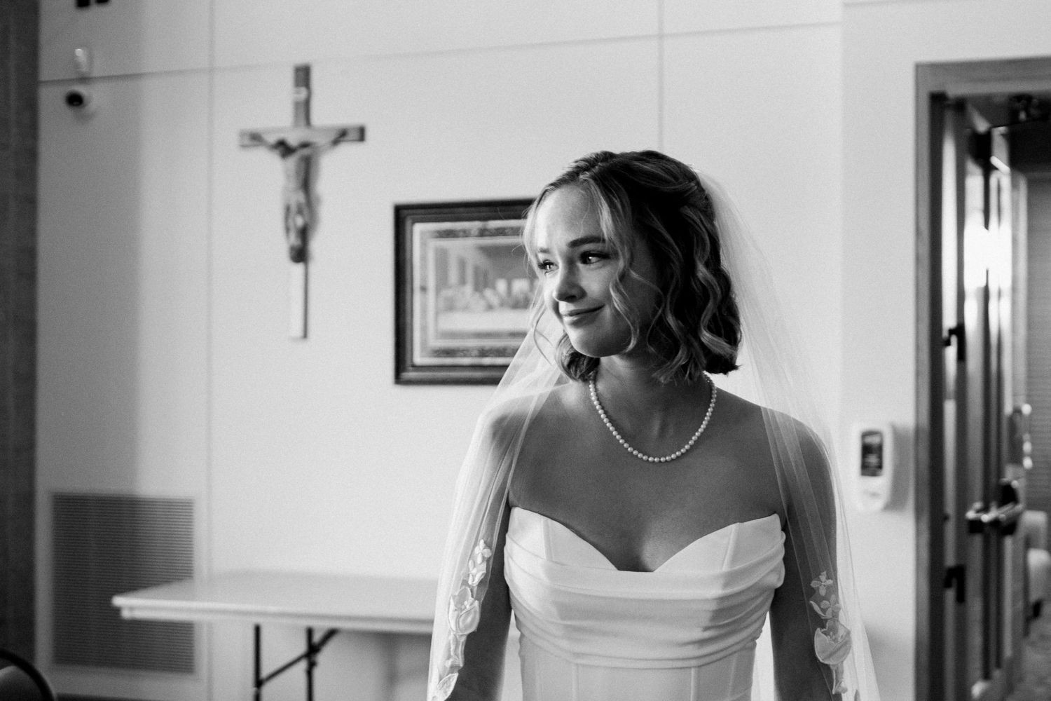 Black and white photo of a smiling bride in a strapless gown and veil, with a crucifix on the wall behind her.