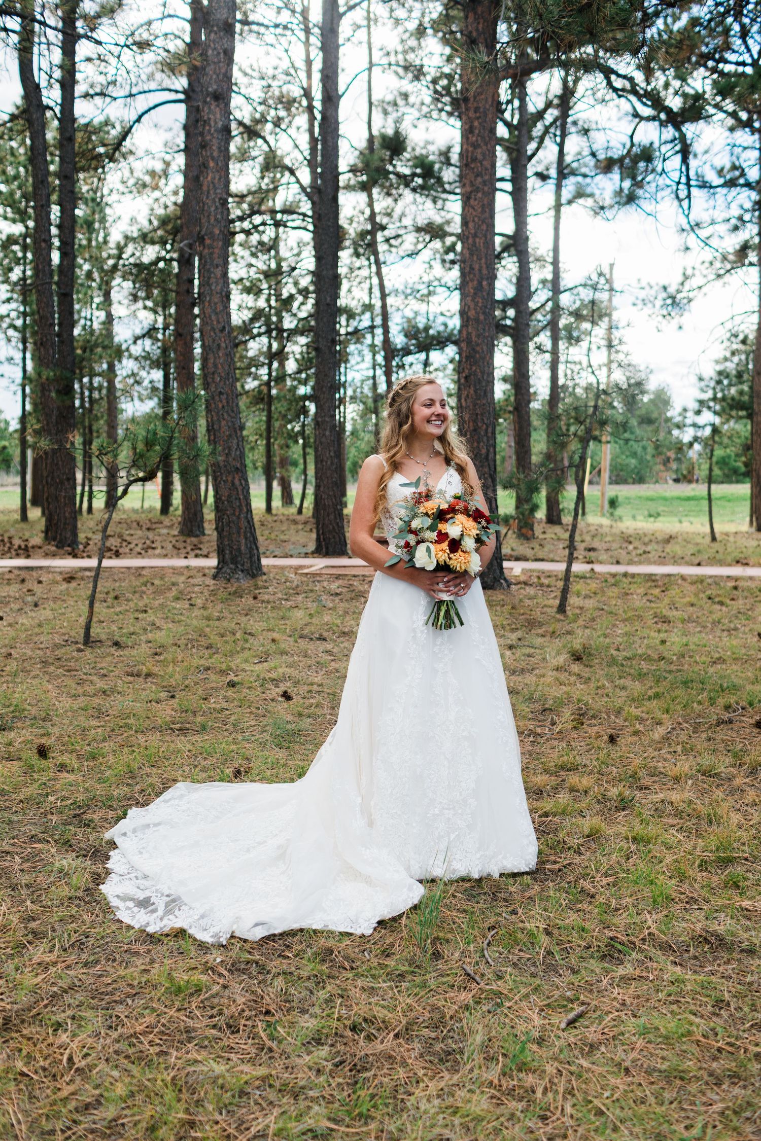 Smiling bride in white lace gown holding colorful bouquet standing among tall pine trees outdoors.