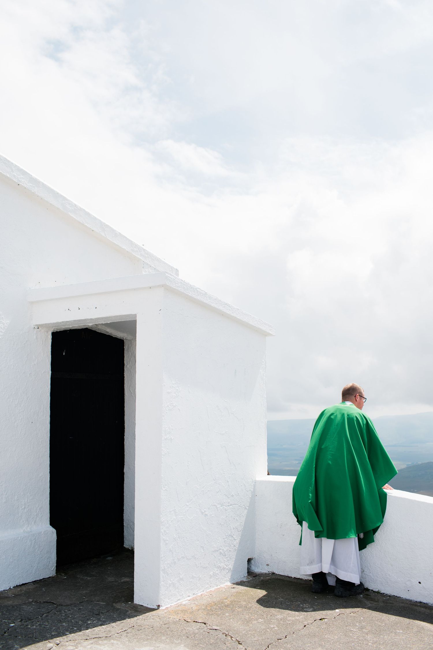 A Catholic priest in green vestments stands on a white church rooftop overlooking cloudy skies.