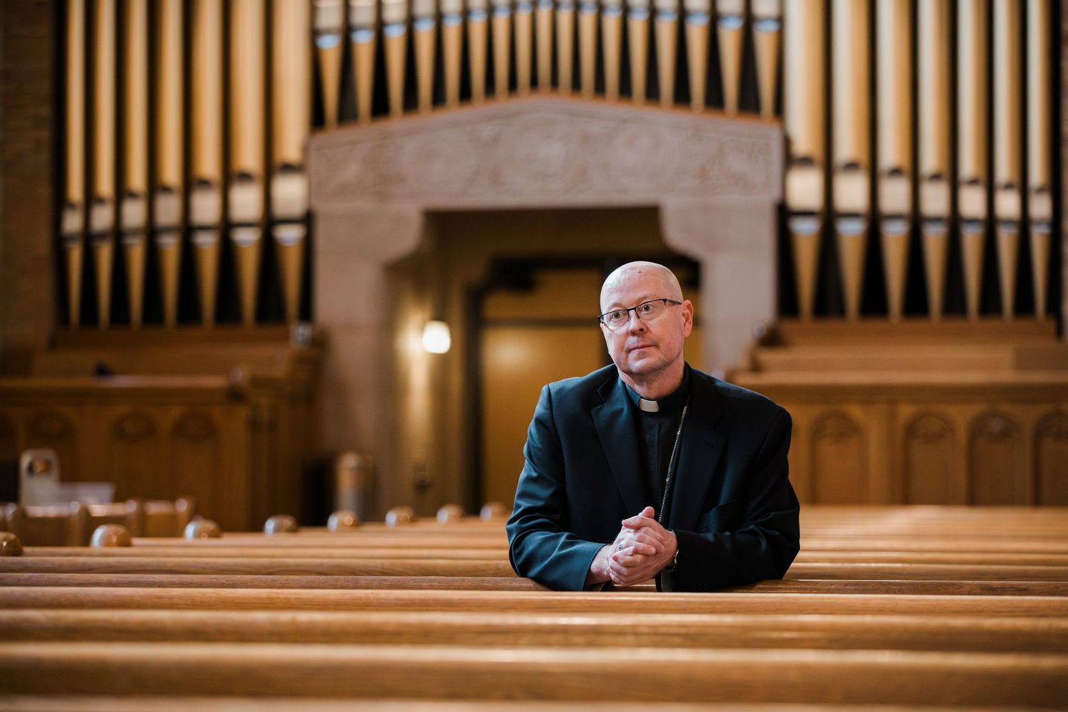 Catholic bishop in black vestment sitting in church pew with large pipe organ in background.