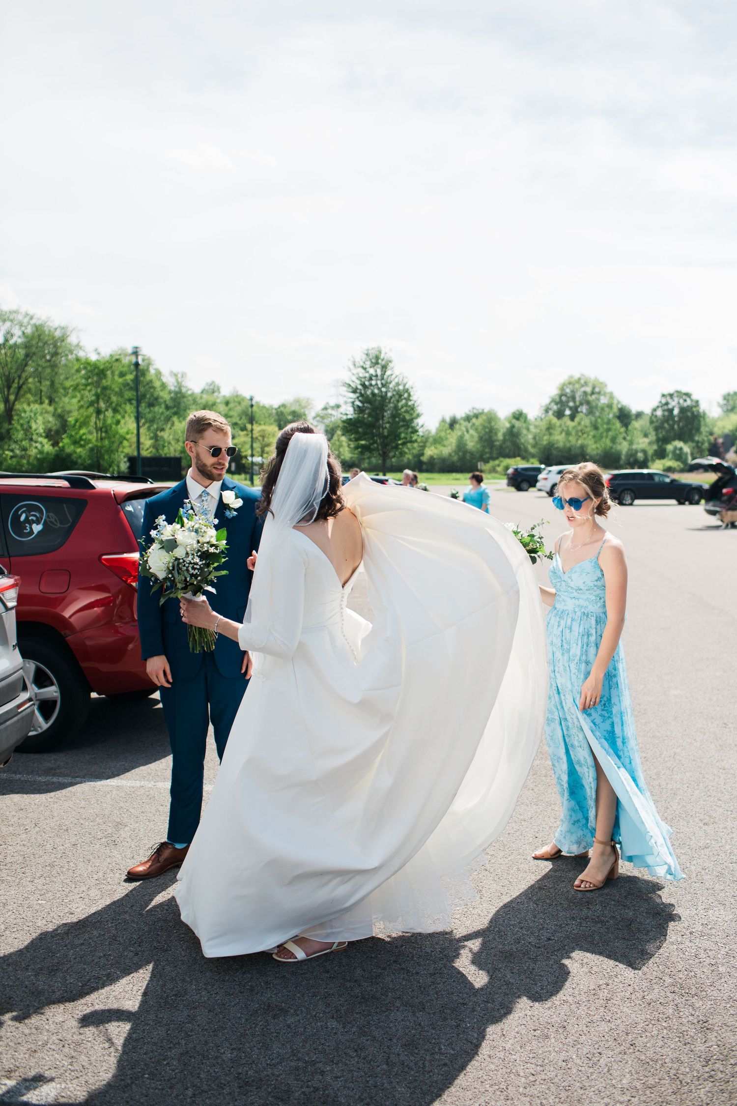 Bride in white gown and veil with groom in navy suit and bridesmaid in blue dress in sunny parking lot.