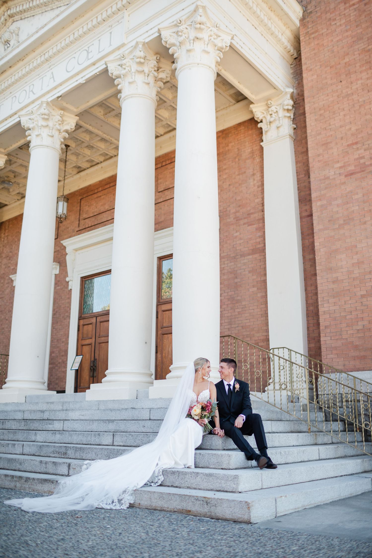 Bride and groom sit on steps of elegant columned church building on their wedding day.