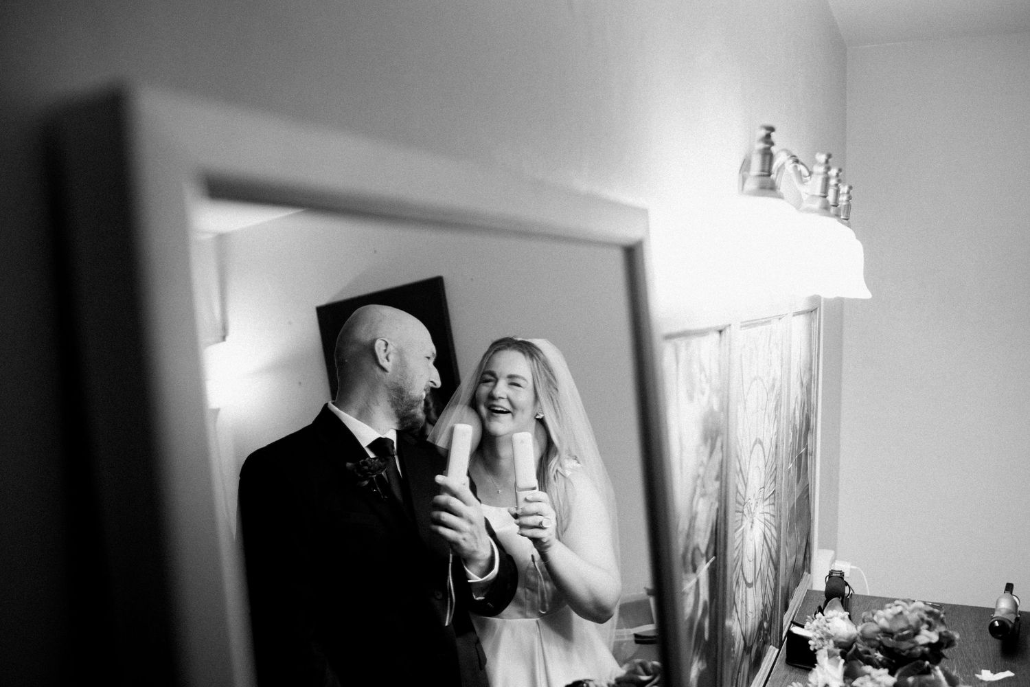 Black and white photo of a laughing bride and groom sharing a joyful moment together reflected in a mirror.