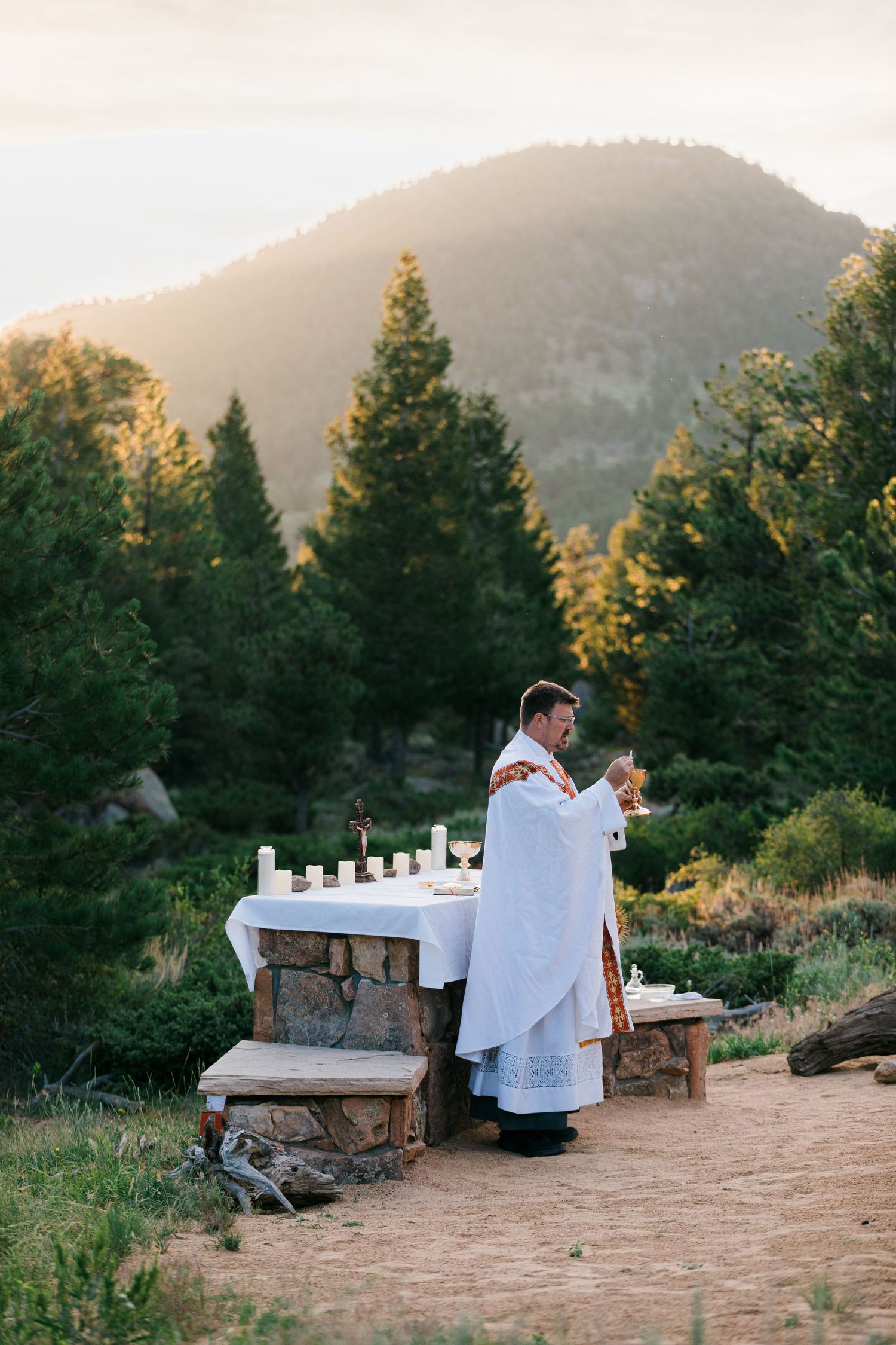 Priest in white vestments performing outdoor Mass at stone altar with mountain and pine trees at sunset.