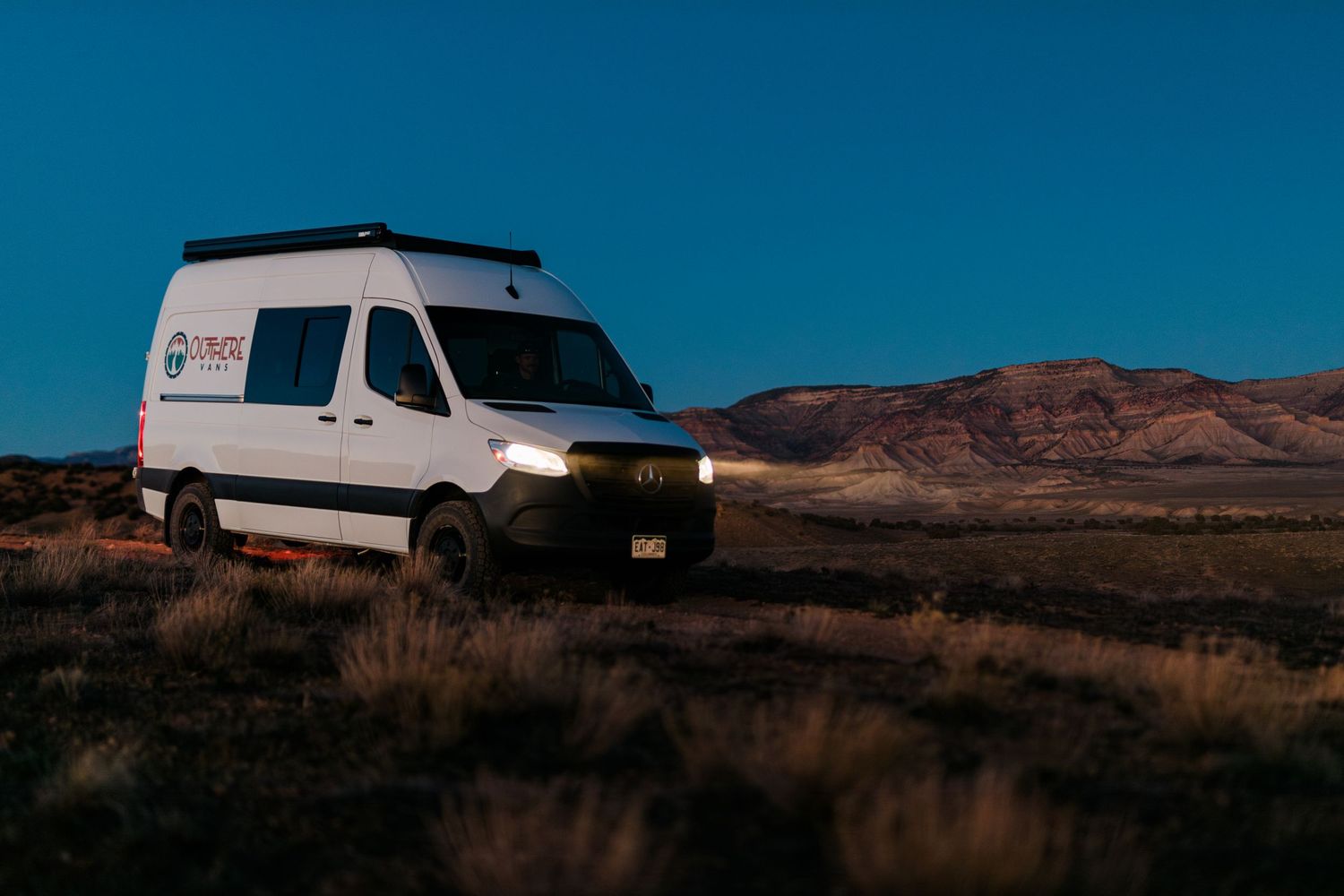 White camper van parked in desert landscape with red mountains and blue sky in background during golden hour.