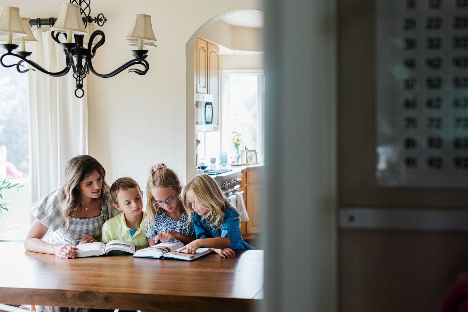 A mother and three children reading books together at a wooden dining table in a bright, cozy home.
