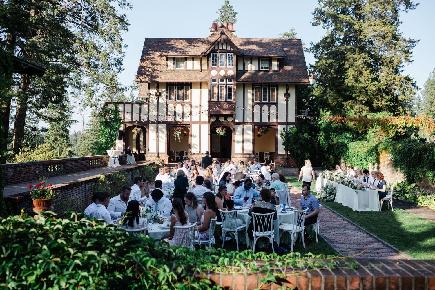 Guests gather for an outdoor wedding reception on the grounds of a Tudor-style mansion at sunset.