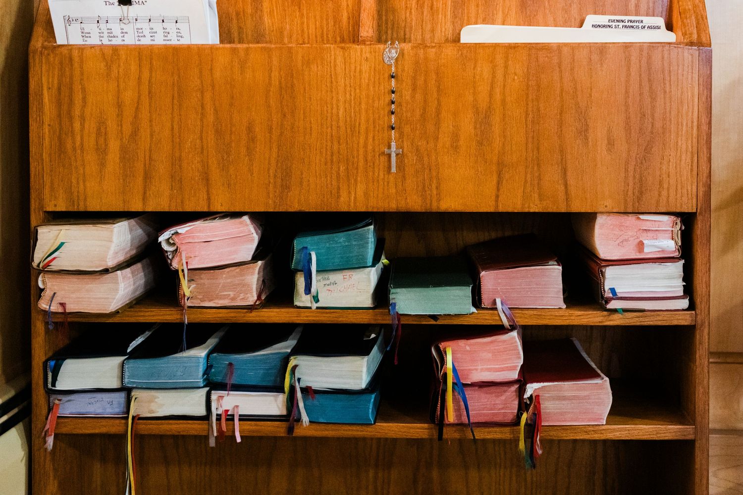 Wooden bookshelf displaying colorful religious hymnals and prayer books with ribbon bookmarks in a church setting.