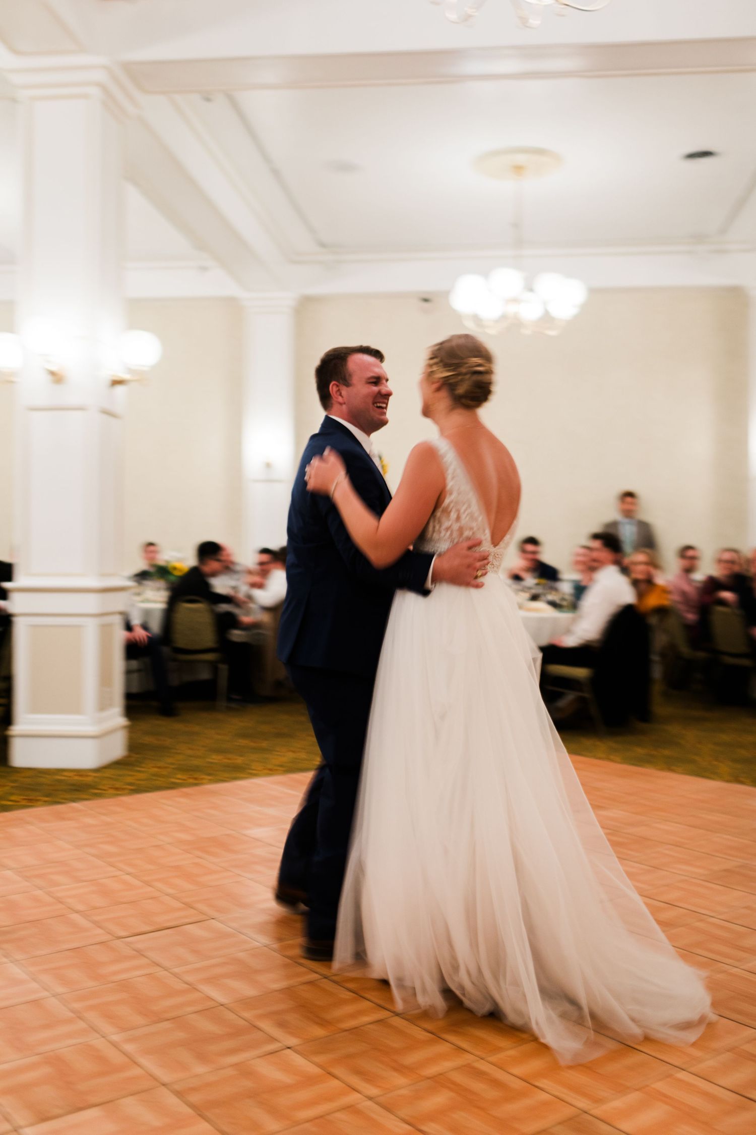 Bride and groom share their first dance in an elegant ballroom with chandeliers and guests seated at tables.
