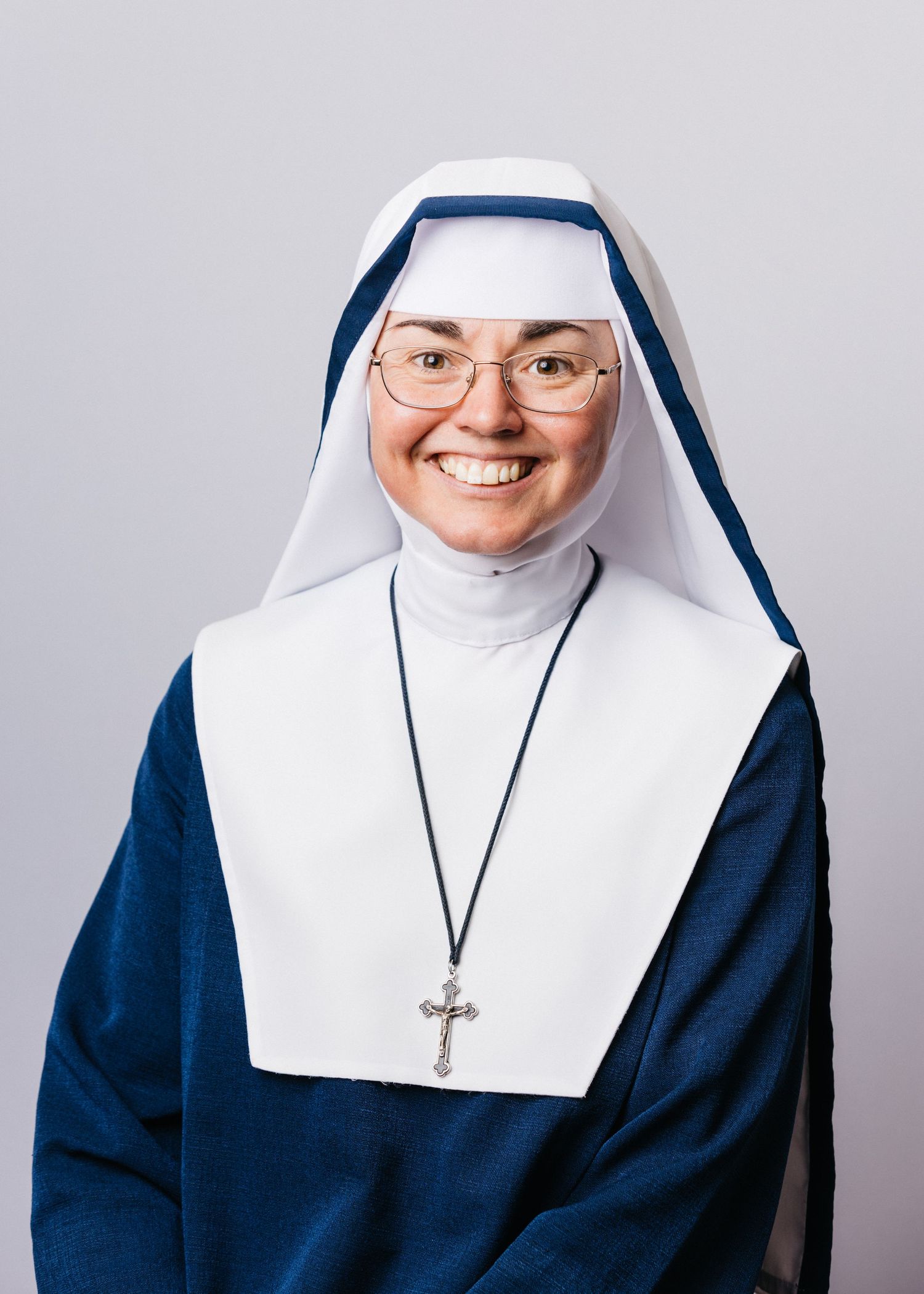 A Catholic nun wearing a traditional black and white habit with glasses and a cross necklace poses for a portrait.