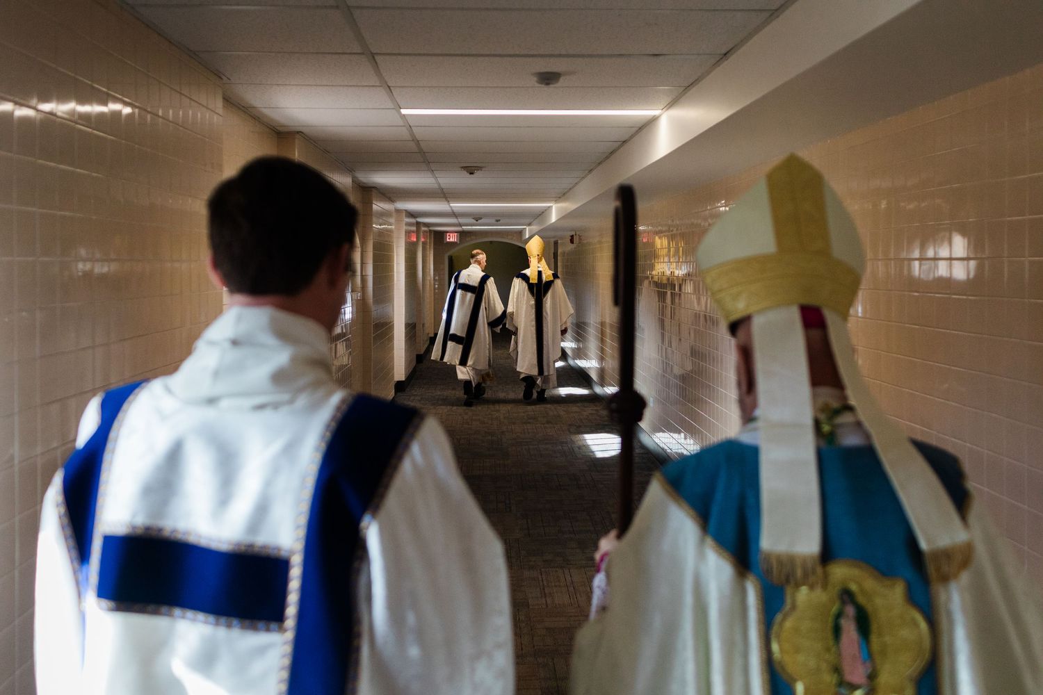 Catholic clergy in white vestments and bishop with mitre walking through institutional hallway.
