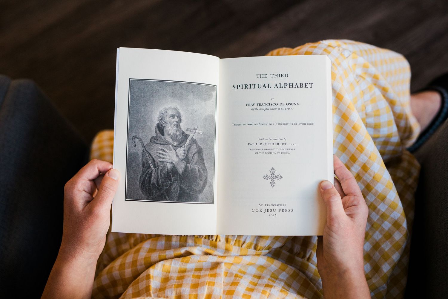 Hands hold open pages of an antique book against a plaid fabric background in warm lighting.