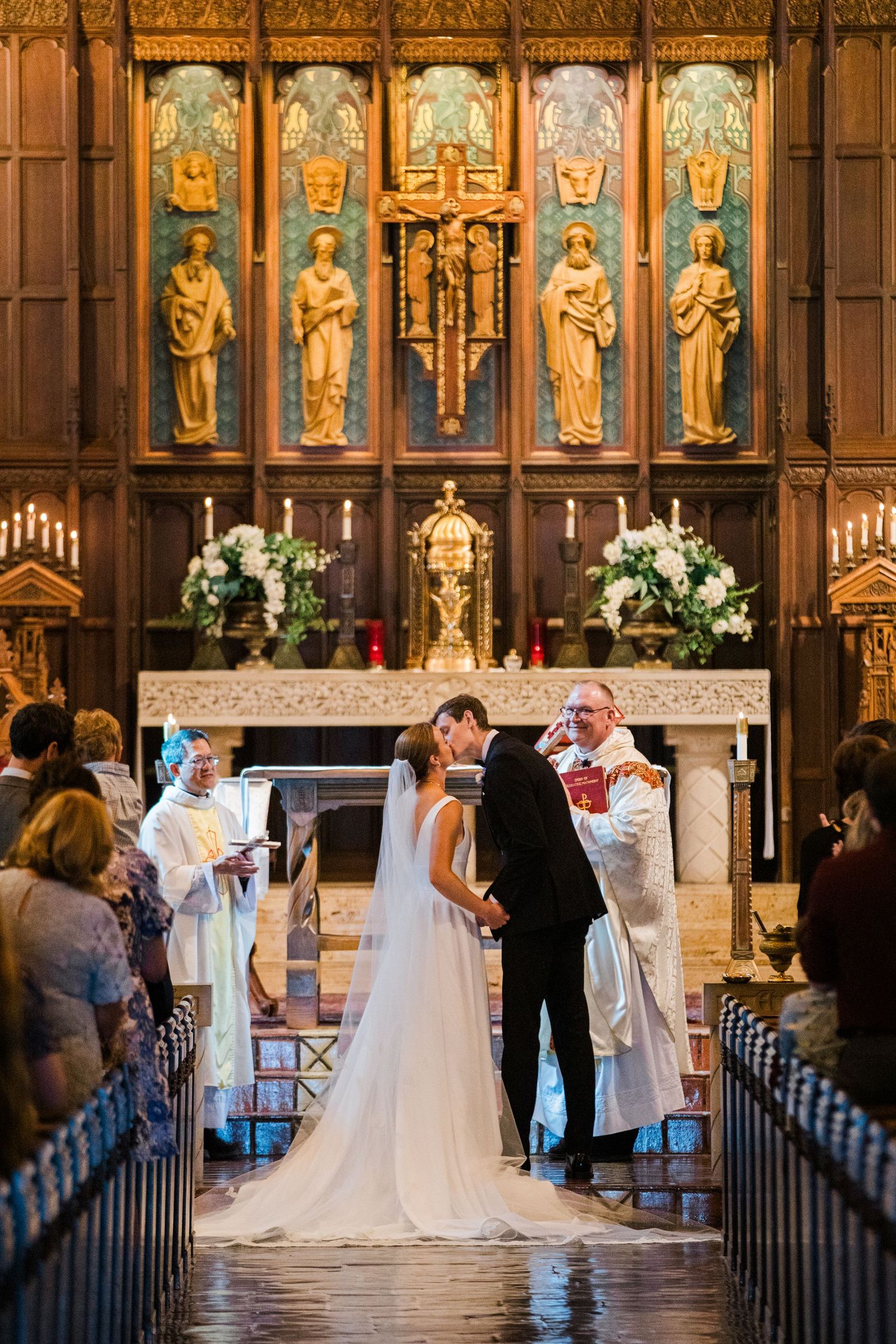 Couple shares first kiss at altar during elegant church wedding ceremony with golden reredos backdrop.