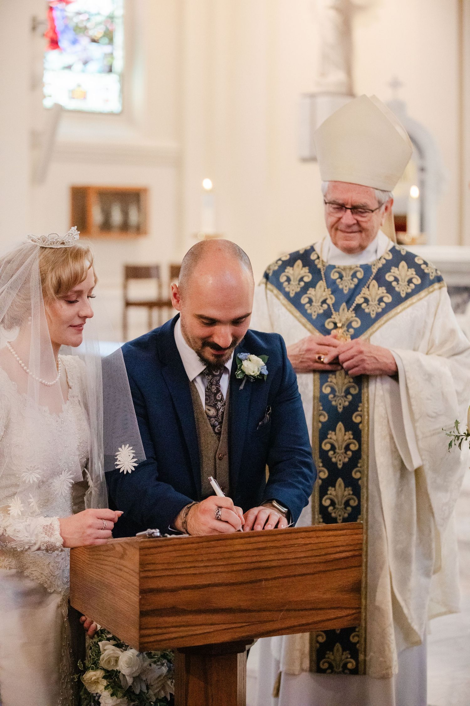 Bride and groom signing wedding registry at church altar with officiating bishop in ornate vestments.