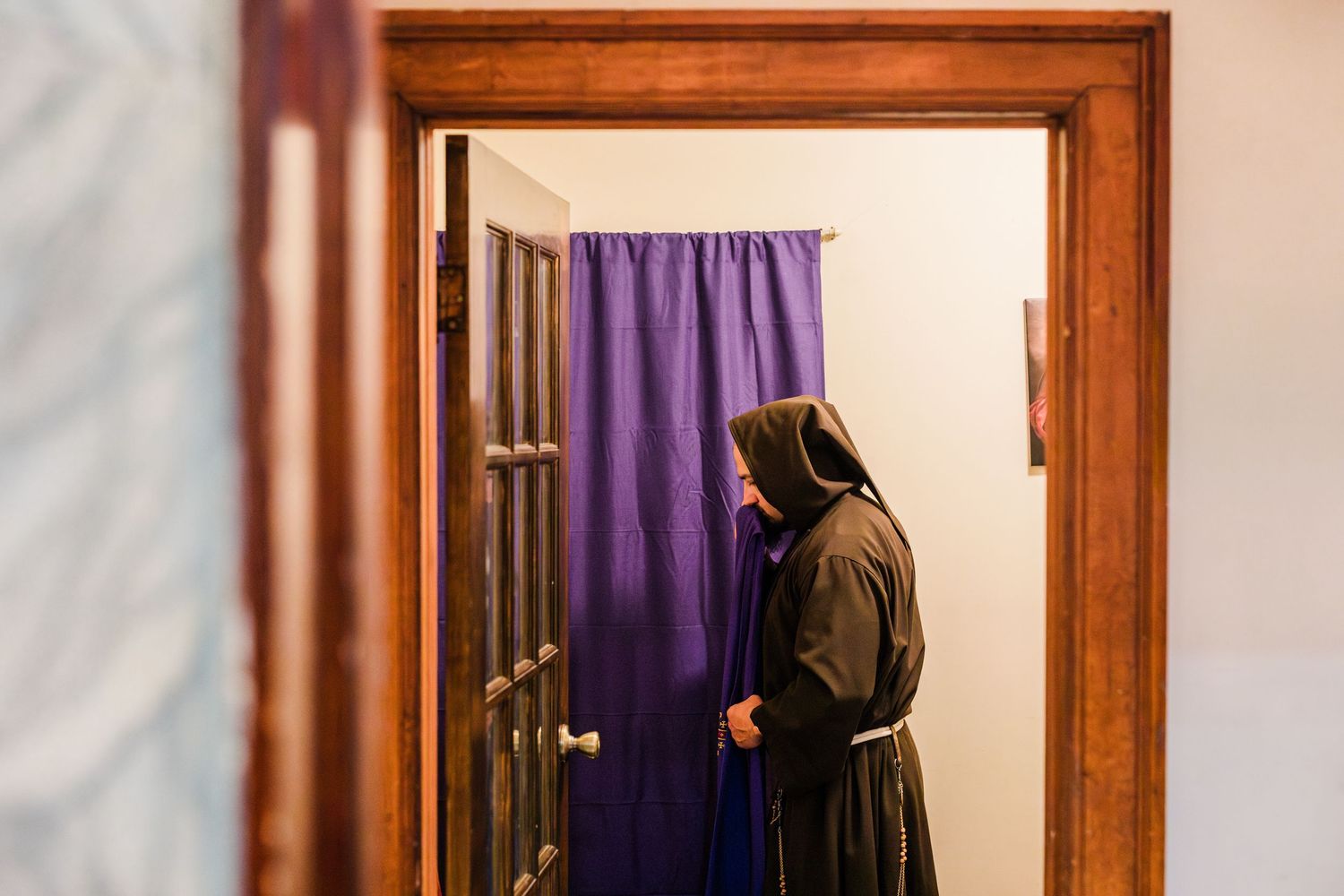A nun in brown habit stands by a wooden door with purple curtain in a serene indoor setting.