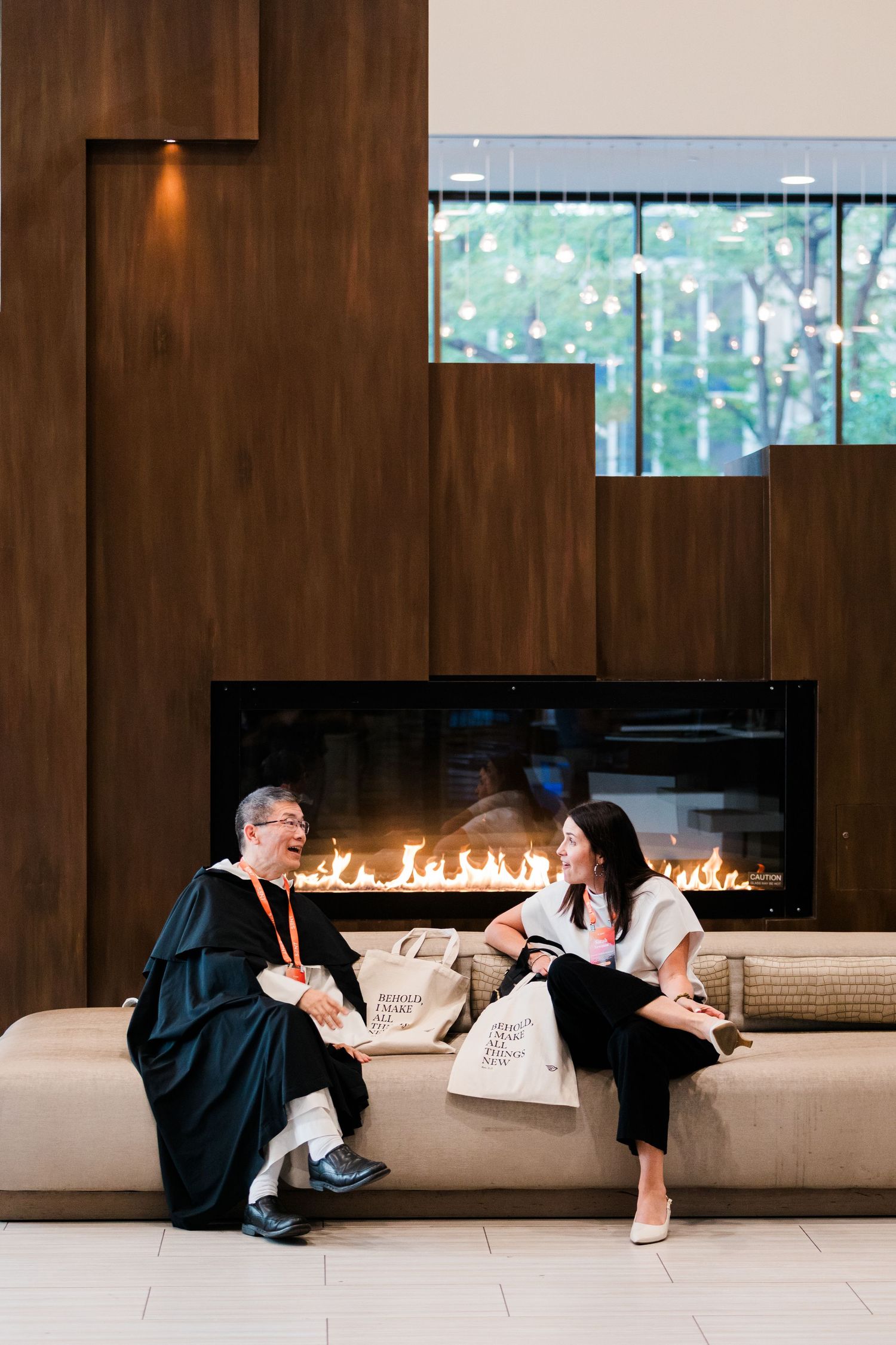 Two people in academic robes chat by a modern fireplace in an elegant wood-paneled lobby during a graduation event.