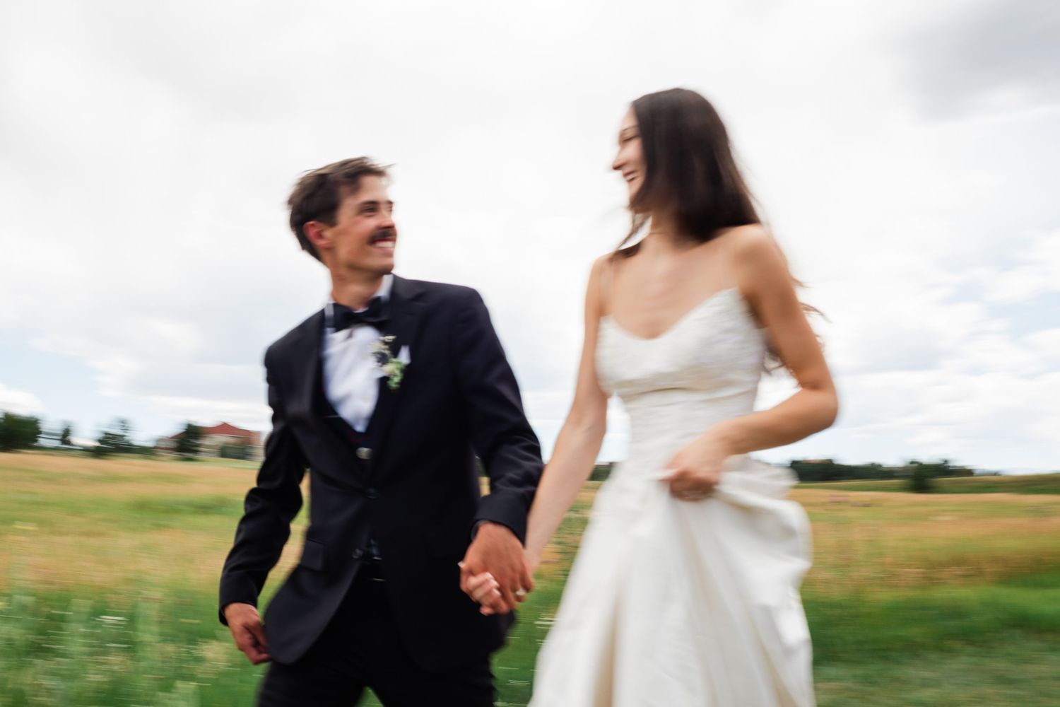 A couple in wedding attire runs playfully through a grassy field on their special day.