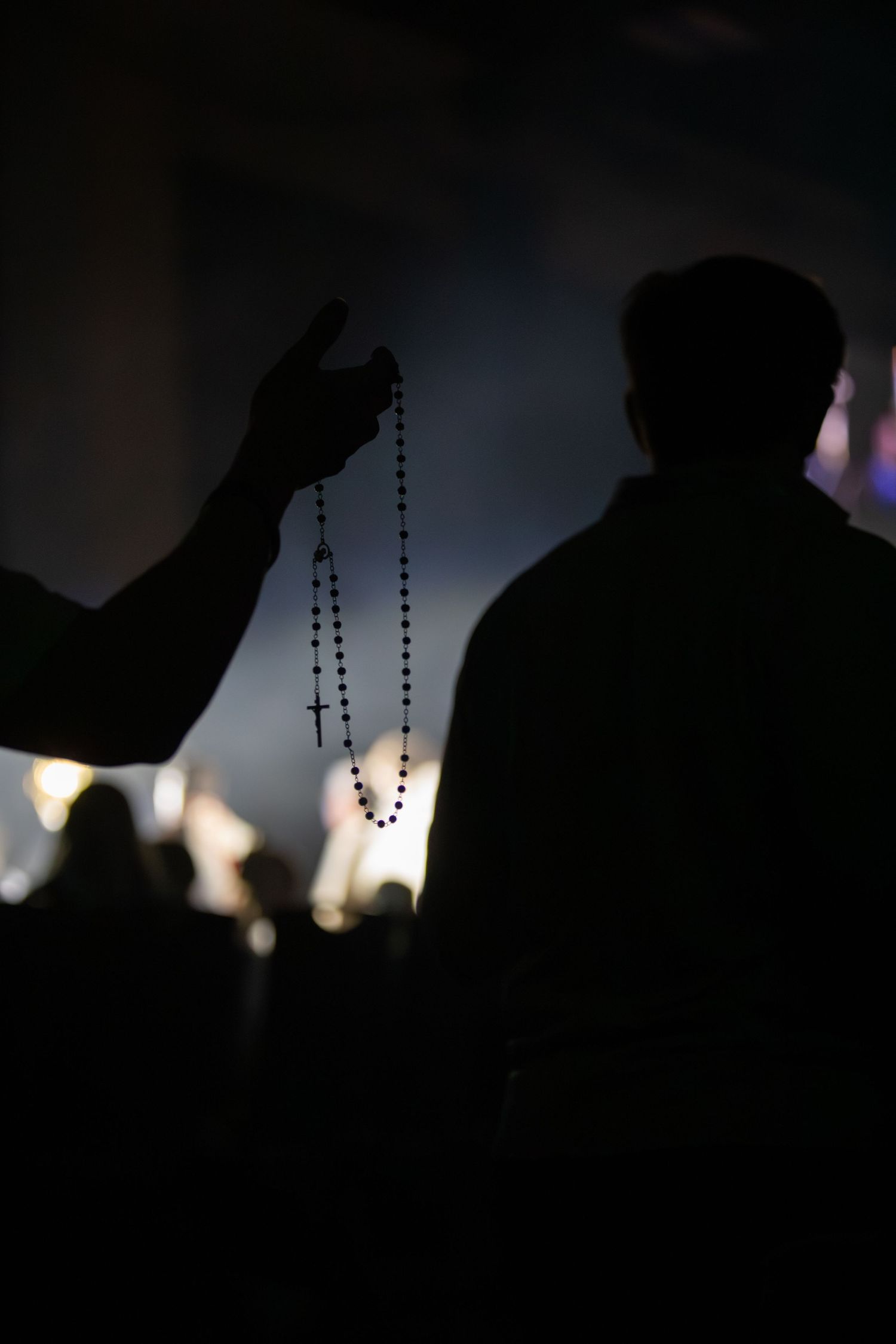 Dark silhouettes of people holding rosary beads against a dim background with candlelight.