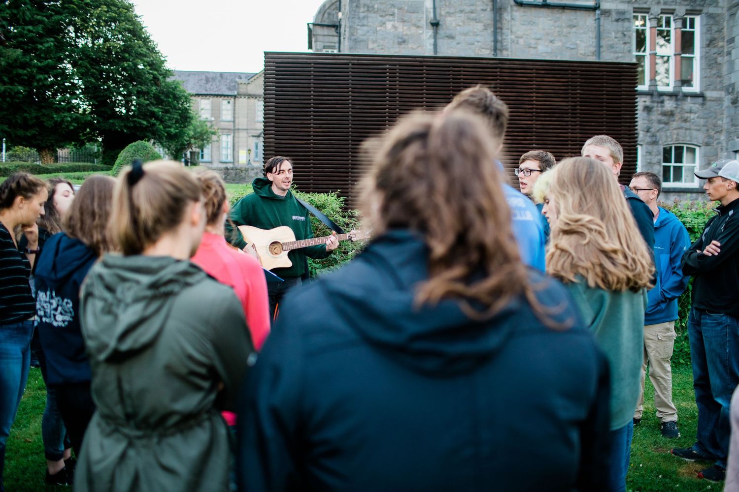 Young people gathered outdoors listening to a guitarist perform during a casual group event.