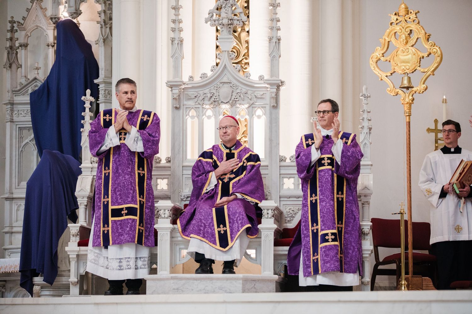 Three Catholic clergy in purple Advent vestments stand at ornate white altar during liturgical celebration.