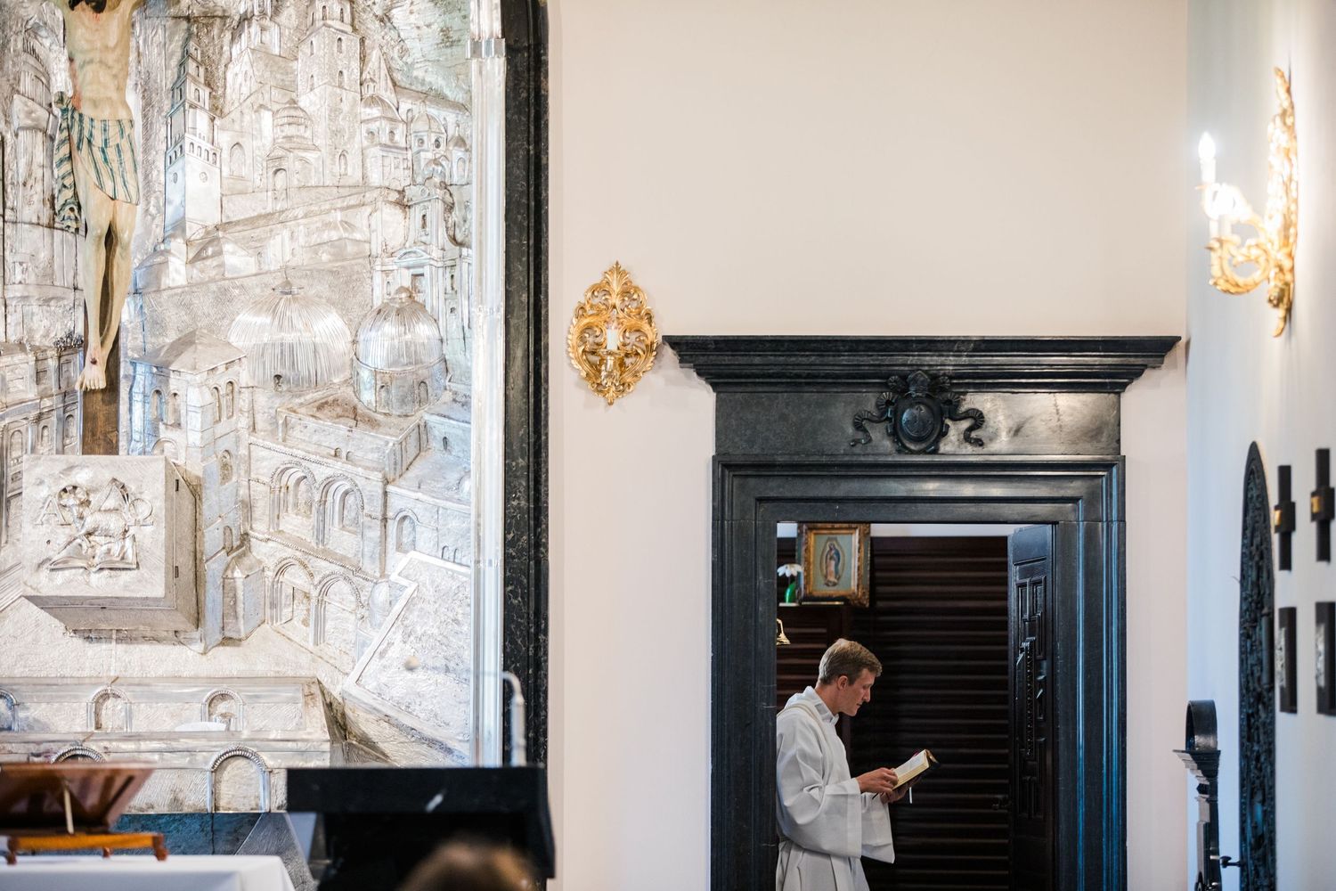 Priest in white robes reads in ornate chapel with gold sconces and detailed silver mural artwork on wall.