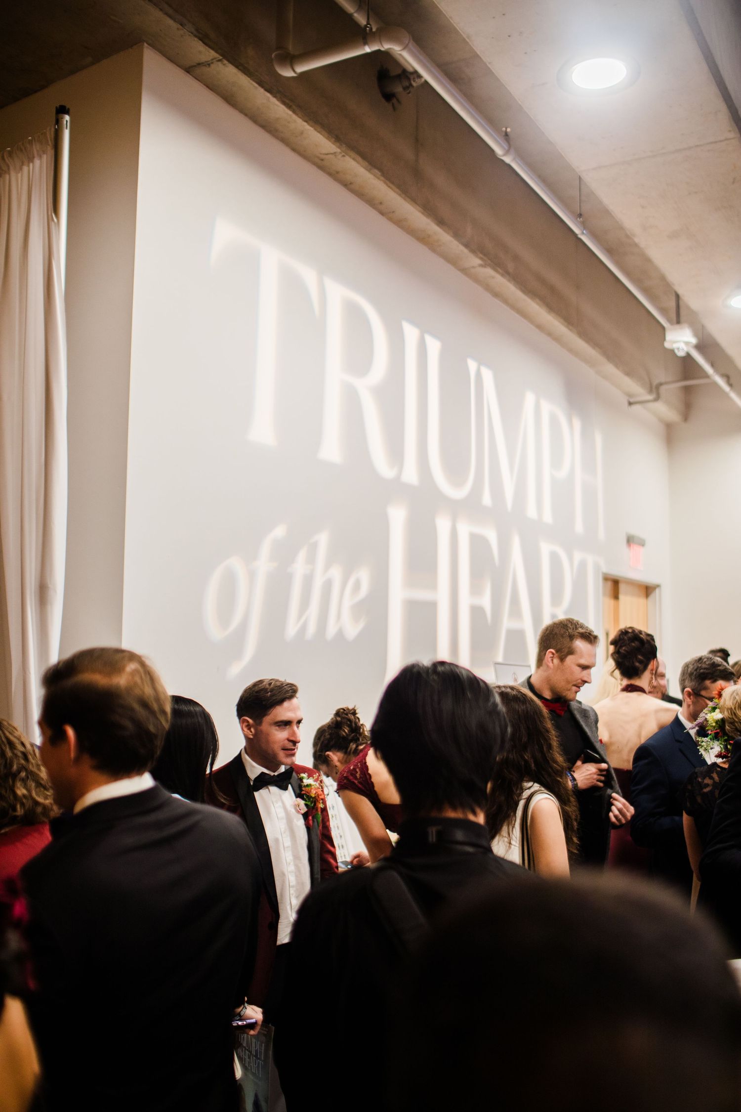 A crowded event space with 'TRIUMPH OF THE HEART' text projected on the wall above formally dressed guests.