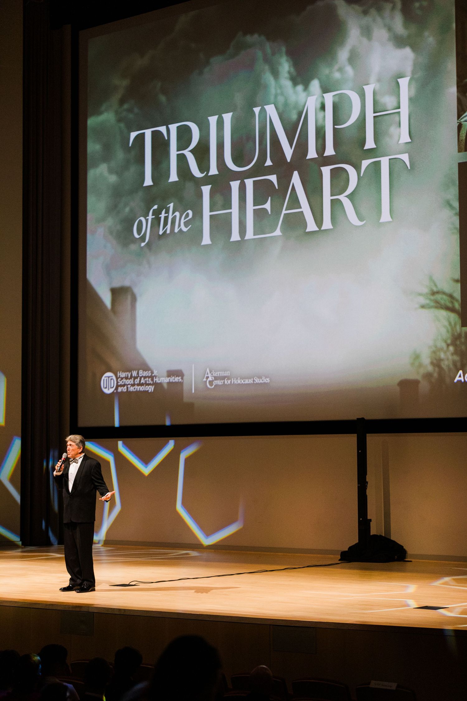 A stage presentation with multiple projection screens showing 'Triumph' text in a dimly lit auditorium setting.