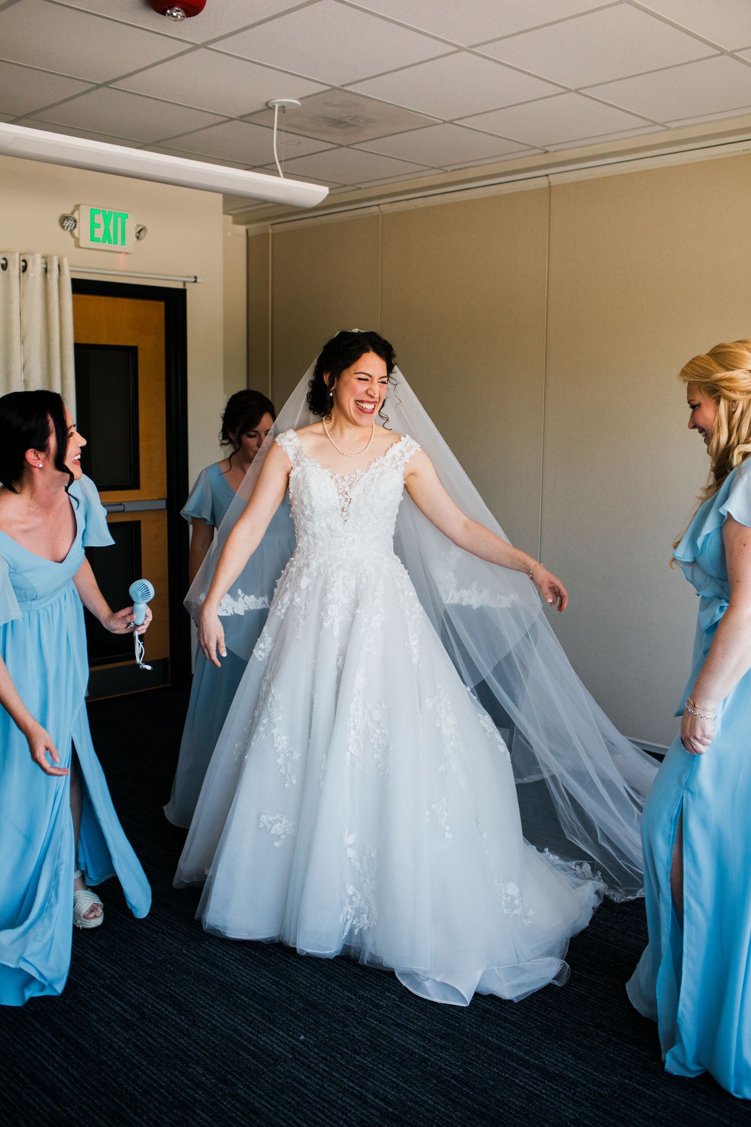 Bride in lace ball gown laughs joyfully surrounded by bridesmaids in light blue dresses during wedding preparations.