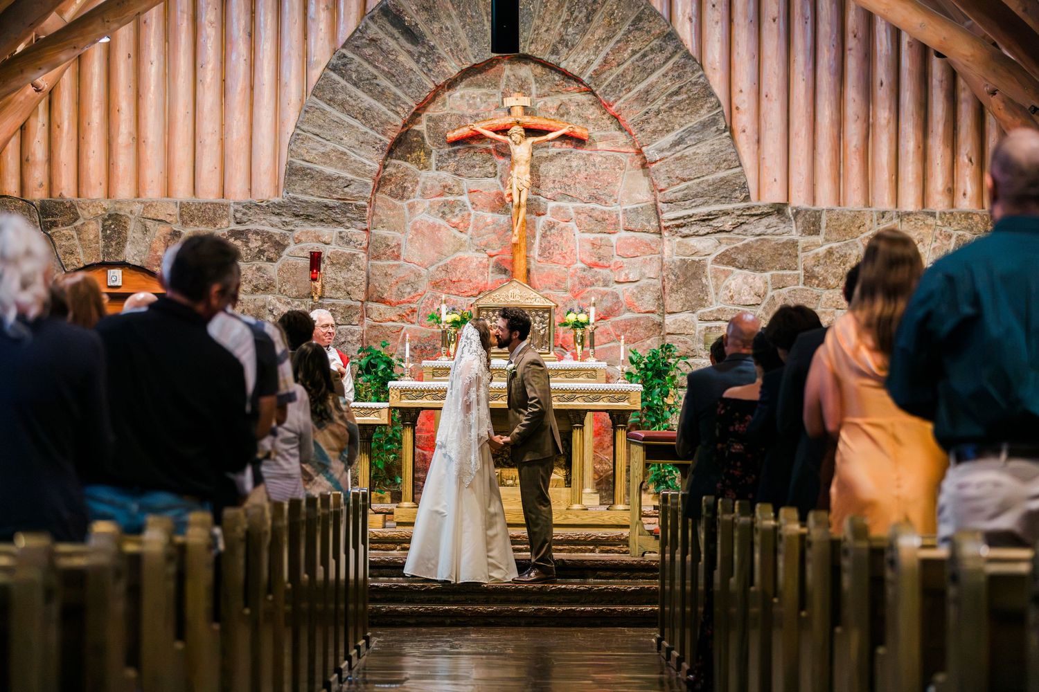 Bride and groom exchange vows at a rustic stone church altar adorned with a crucifix, surrounded by seated wedding guests.