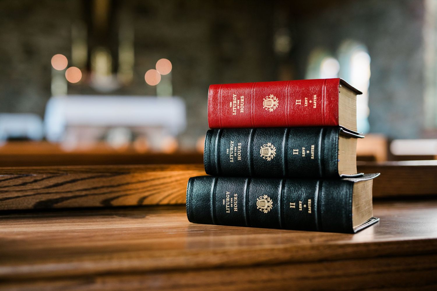 Stack of three Liturgy of the Hours volumes in red and black on a wooden church pew.