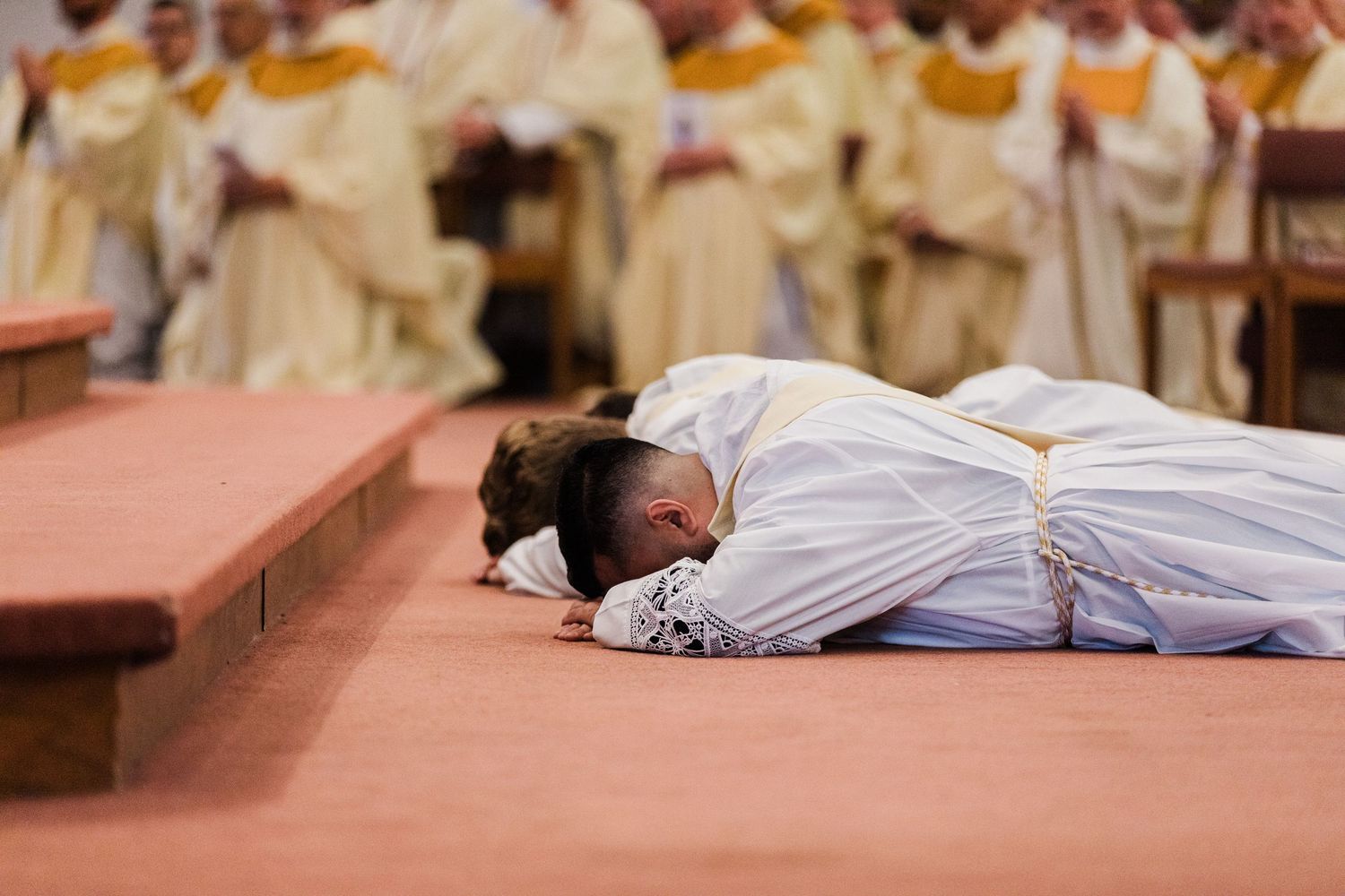 Catholic priest prostrating during ordination ceremony with clergy in white vestments in background.