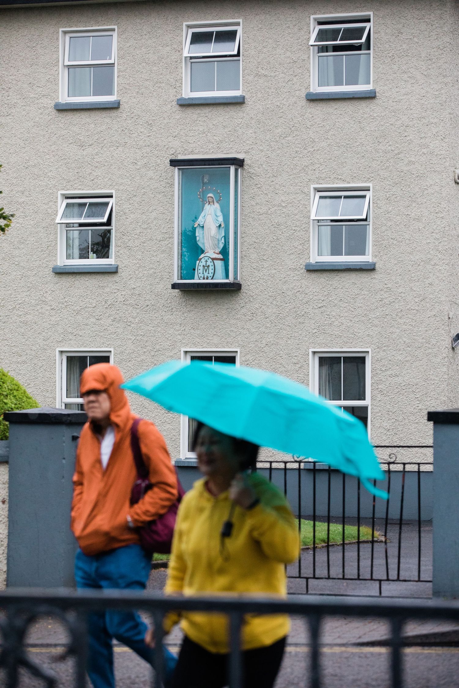 People with umbrellas walking in the rain past apartment buildings on a grey day.