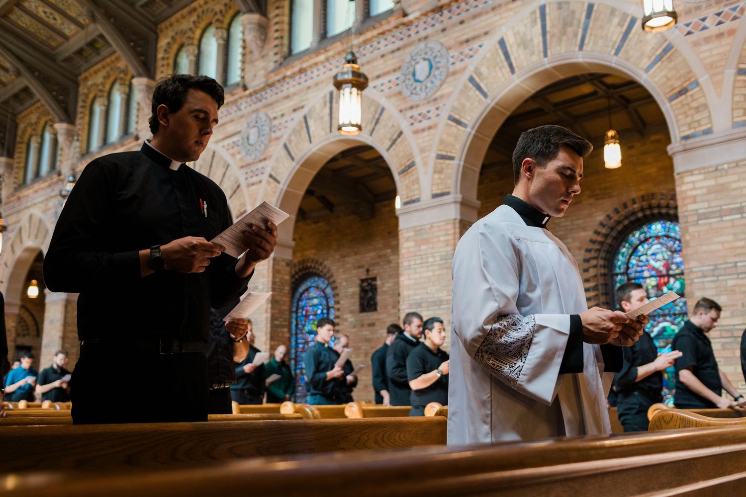 Two clergy members read from papers during a church service in an ornate cathedral with arched stonework and stained glass.
