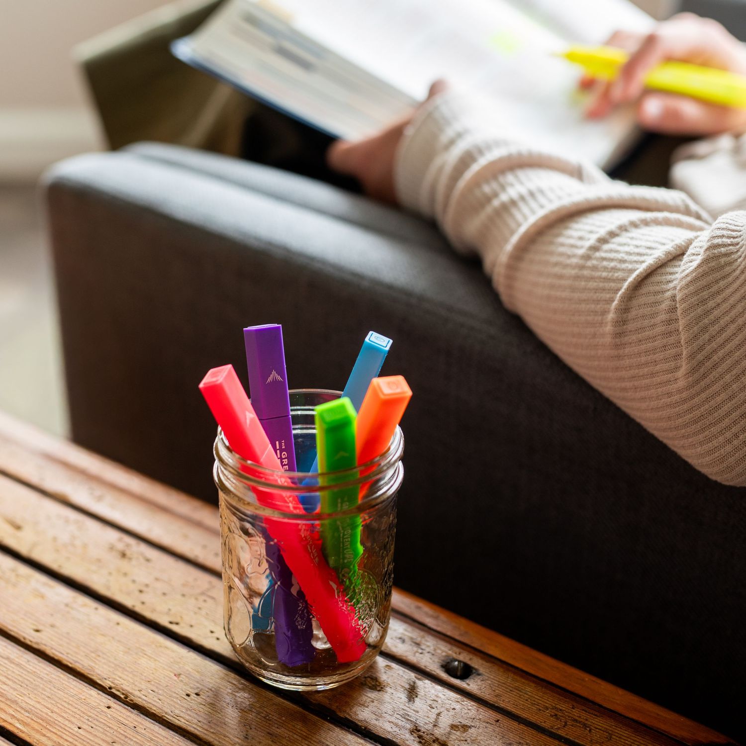 Colorful marker pens arranged in a glass jar on a wooden table next to someone working on a laptop.