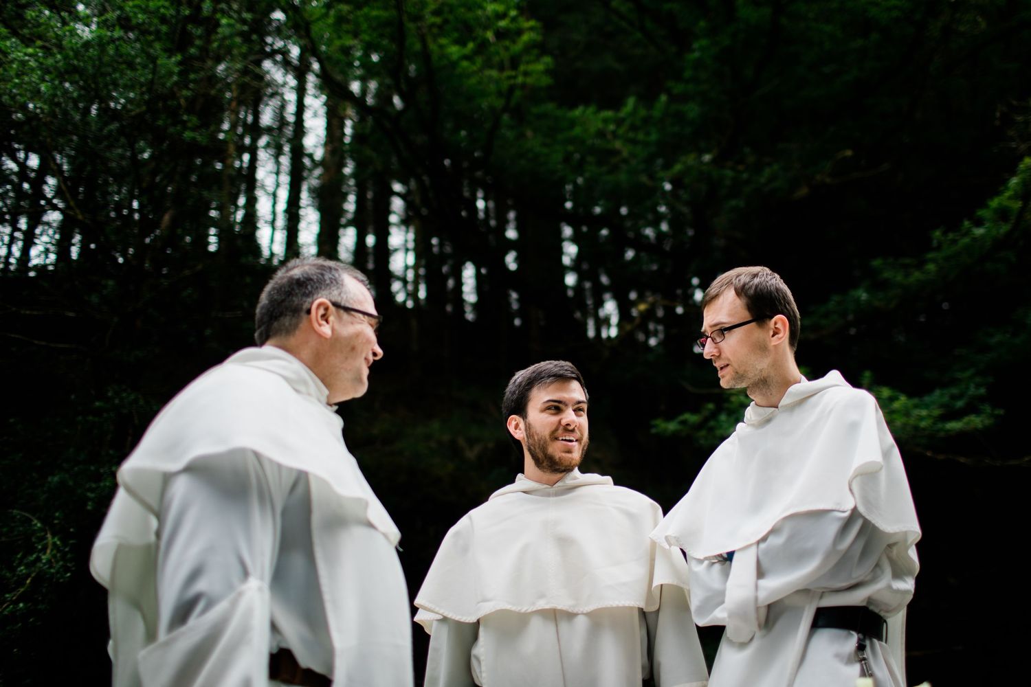 Three Catholic monks in white robes and scapulars engage in conversation outdoors with lush green trees in the background.