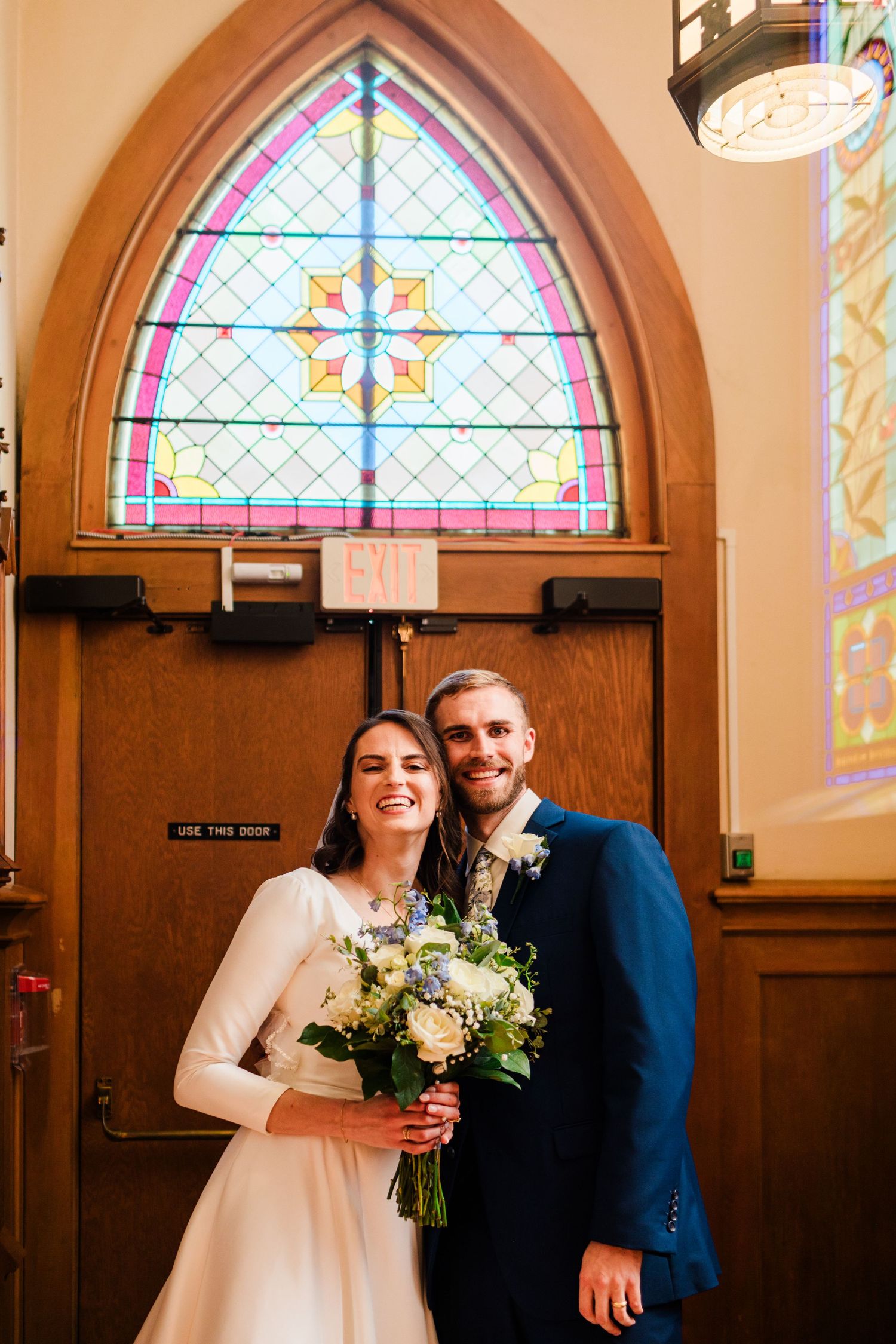 Smiling bride and groom pose together in front of stained glass church windows, bride holding white and blue bouquet.