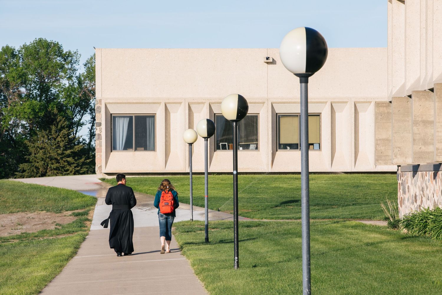 Students walk along a curved concrete path between grassy areas and modern buildings on a college campus.