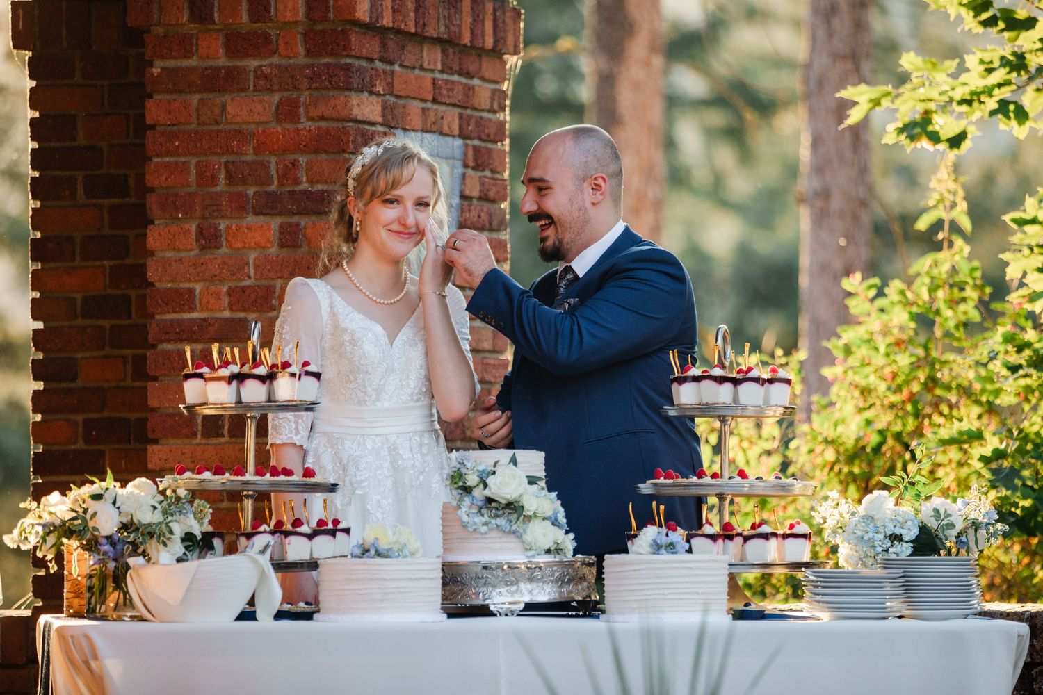 Bride and groom cutting wedding cake outdoors at dessert table with cupcakes and floral arrangements.