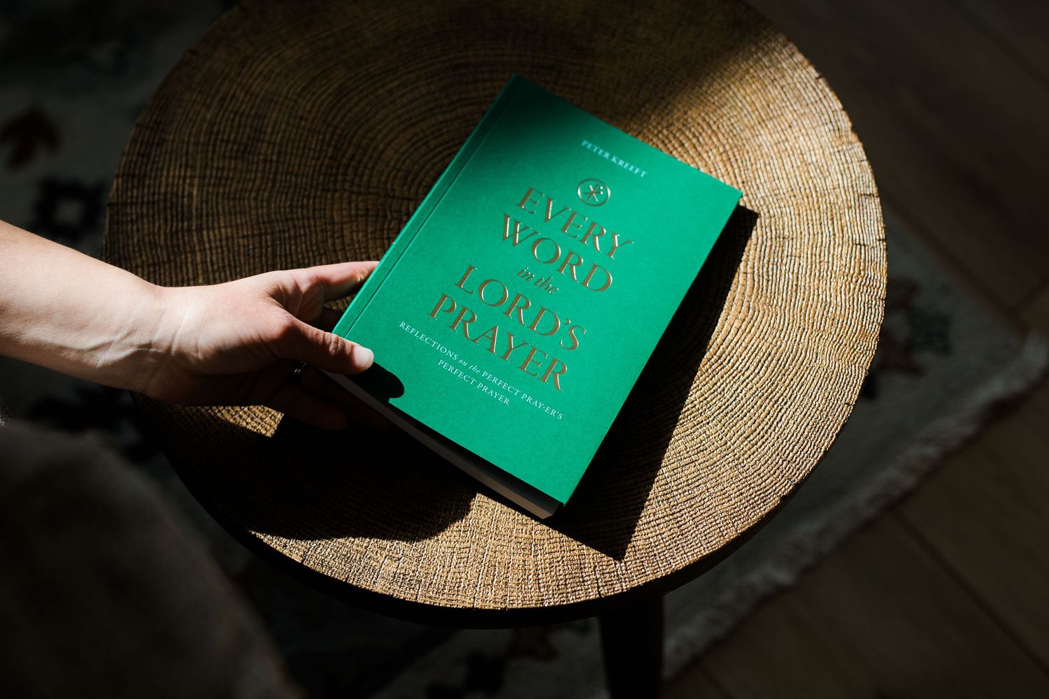 Hand holding a green hardcover Lord's Prayer book on a woven straw hat in natural sunlight.
