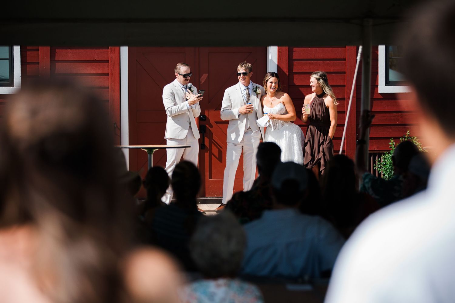 Outdoor wedding ceremony with couple and officiant standing before red barn, guests seated in foreground on sunny day.
