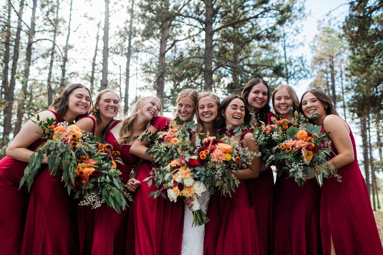 Bride in white gown with bridesmaids in red dresses holding colorful bouquets, smiling outdoors among pine trees.