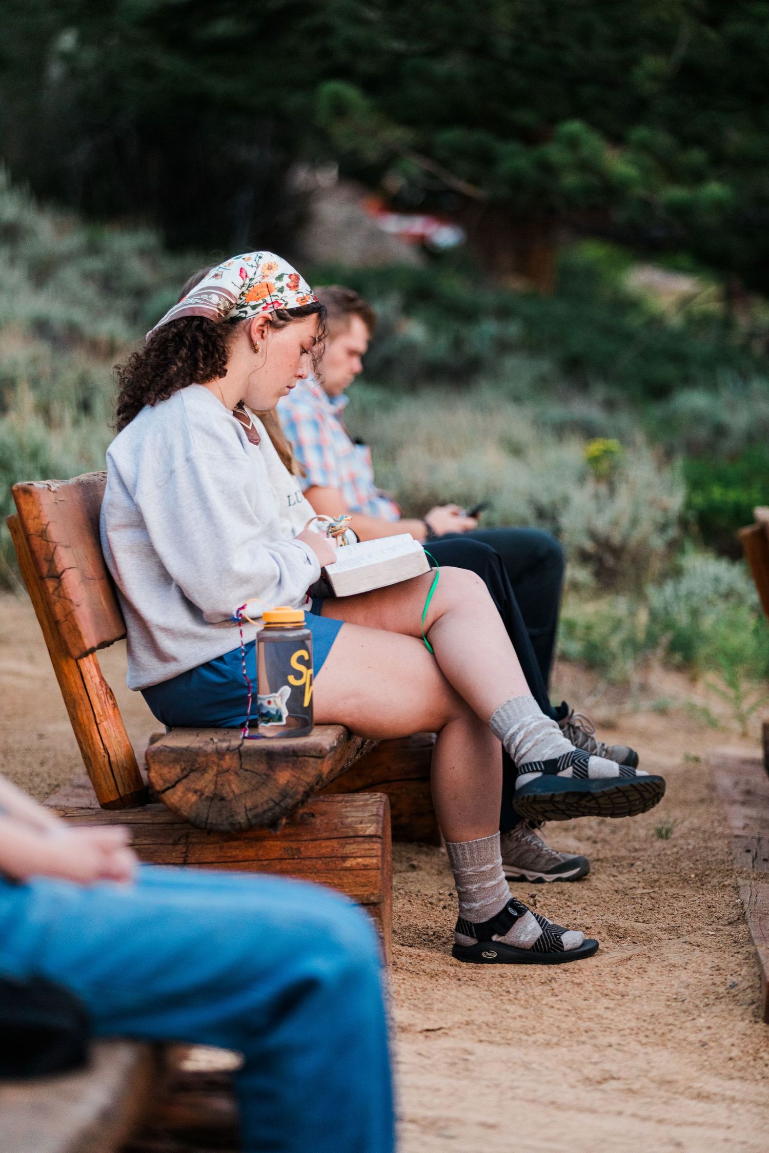 Young woman reading a book on a wooden bench outdoors at a campsite, wearing denim shorts and a floral headband.