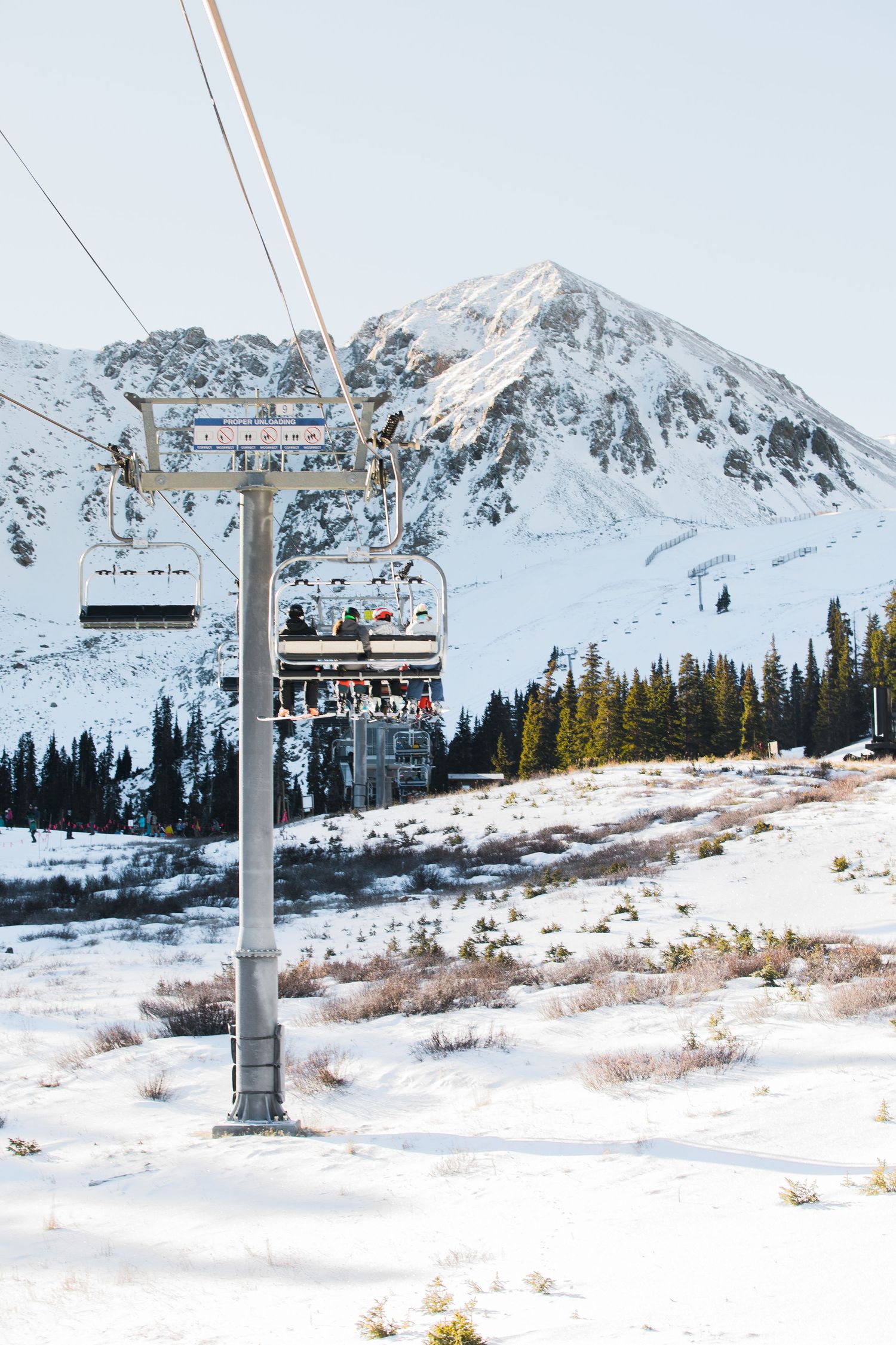 Snow-covered ski lift chairs transport riders up a mountain slope with evergreen trees and dramatic snowy peaks in view.