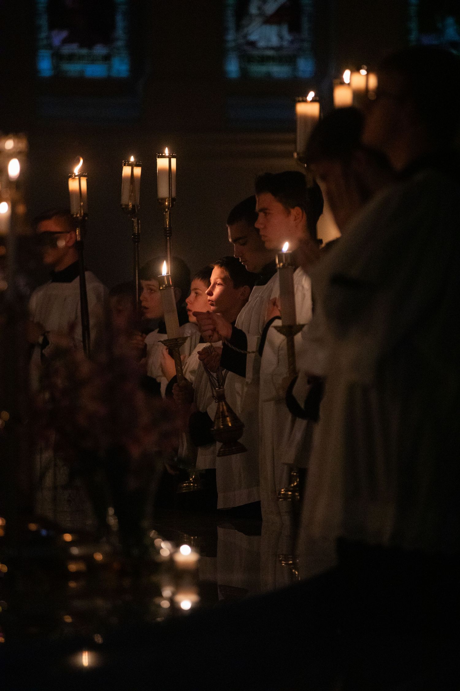 Candlelit procession of clergy members in white robes holding glowing candles in a dark church.