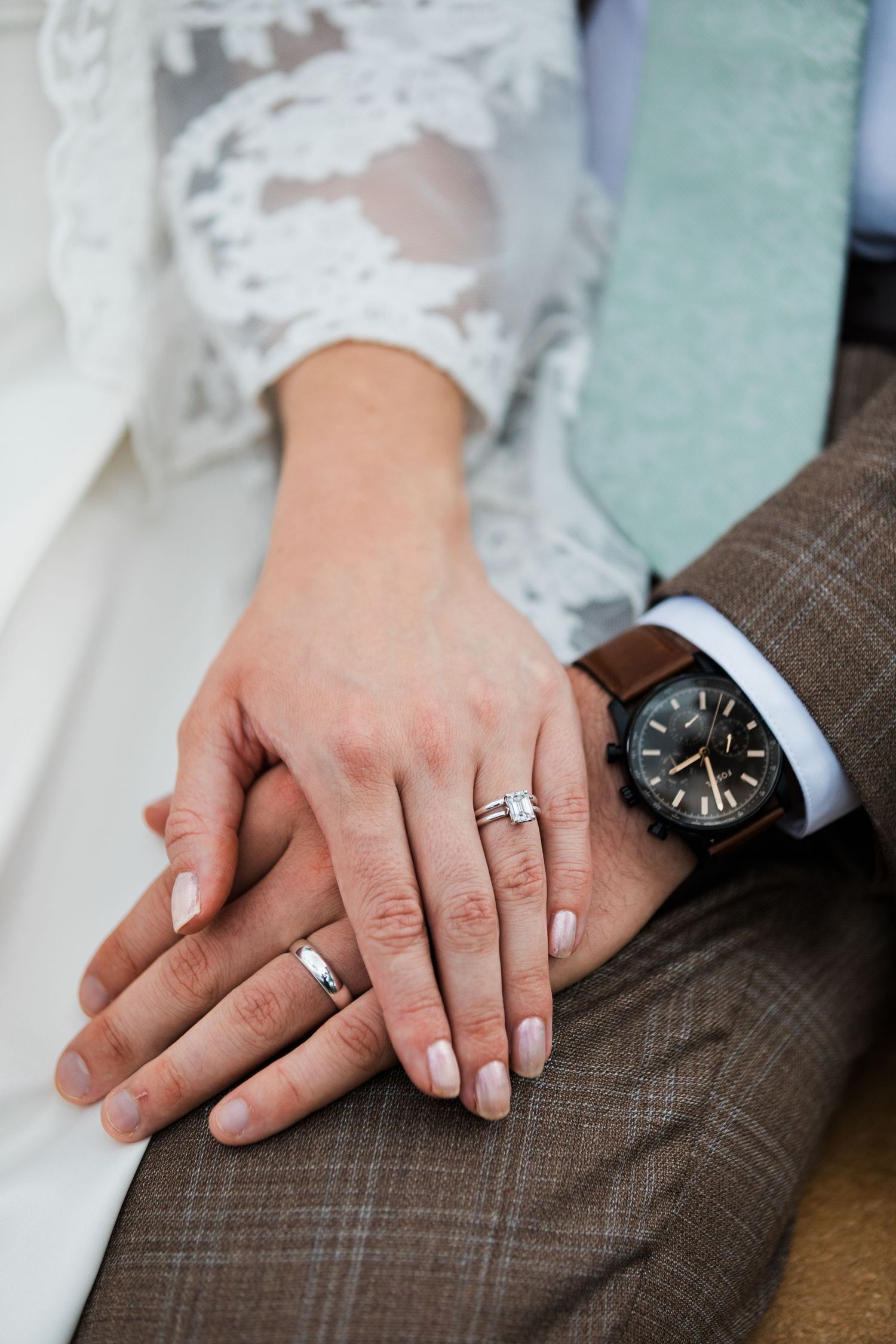Newlyweds displaying wedding rings and engagement ring on joined hands, bride in lace dress, groom in suit with watch.