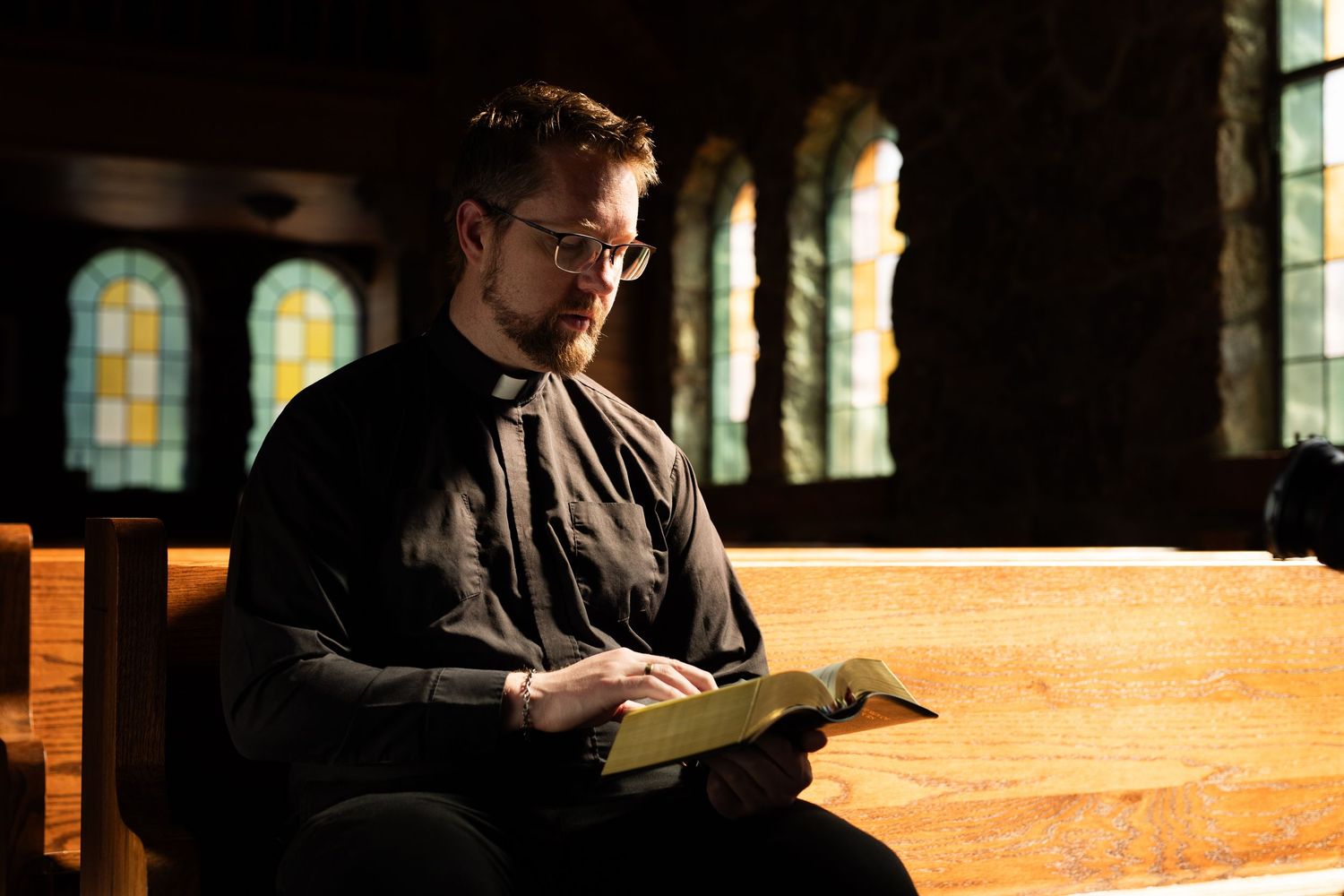 A priest in black clerical attire reads The Liturgy of the Hours in a church pew with stained glass windows.